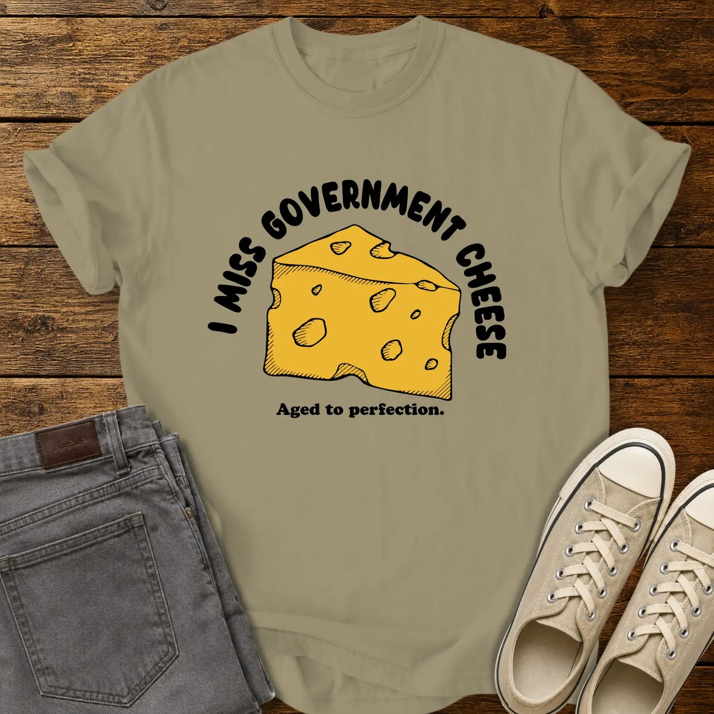 khaki T-shirt mockup featuring a hand-drawn wedge of yellow cheese with the phrase “I MISS GOVERNMENT CHEESE” arched above and “Aged to perfection.” below. The shirt is displayed on a rustic wood background alongside folded gray jeans and cream-colored canvas sneakers.