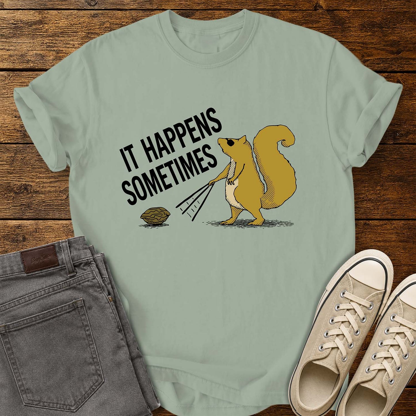 Bay green T-shirt featuring a humorous retro illustration of a squirrel using a rake to pull a walnut toward itself. Bold black text angled above the artwork reads, “IT HAPPENS SOMETIMES.” The shirt is displayed on a rustic wood plank background with folded gray jeans on the left and cream canvas sneakers on the right.
