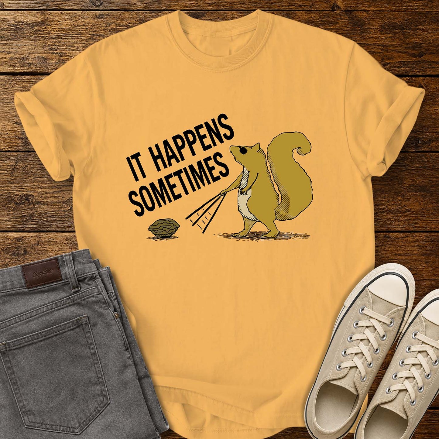 citrus yellow T-shirt featuring a humorous retro illustration of a squirrel using a rake to pull a walnut toward itself. Bold black text angled above the artwork reads, “IT HAPPENS SOMETIMES.” The shirt is displayed on a rustic wood plank background with folded gray jeans on the left and cream canvas sneakers on the right.