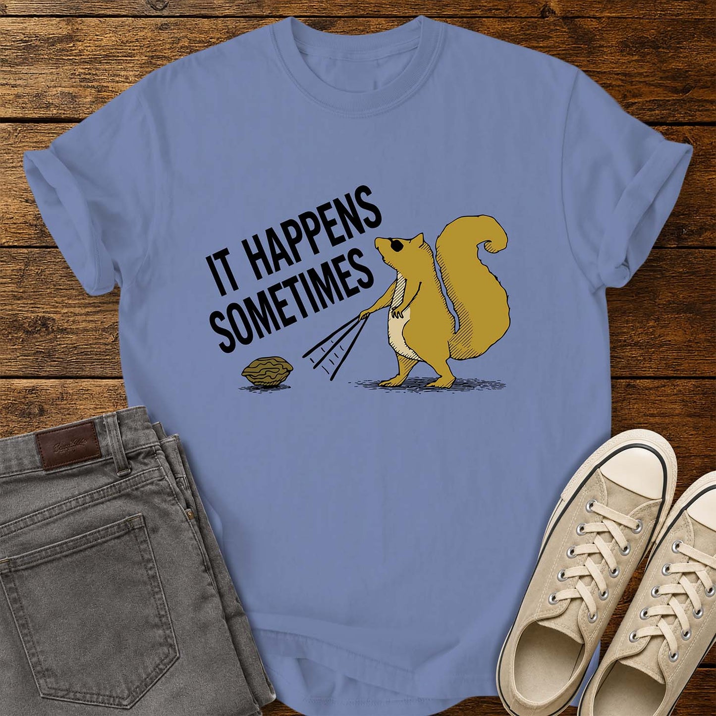 washed denim blue T-shirt featuring a humorous retro illustration of a squirrel using a rake to pull a walnut toward itself. Bold black text angled above the artwork reads, “IT HAPPENS SOMETIMES.” The shirt is displayed on a rustic wood plank background with folded gray jeans on the left and cream canvas sneakers on the right.