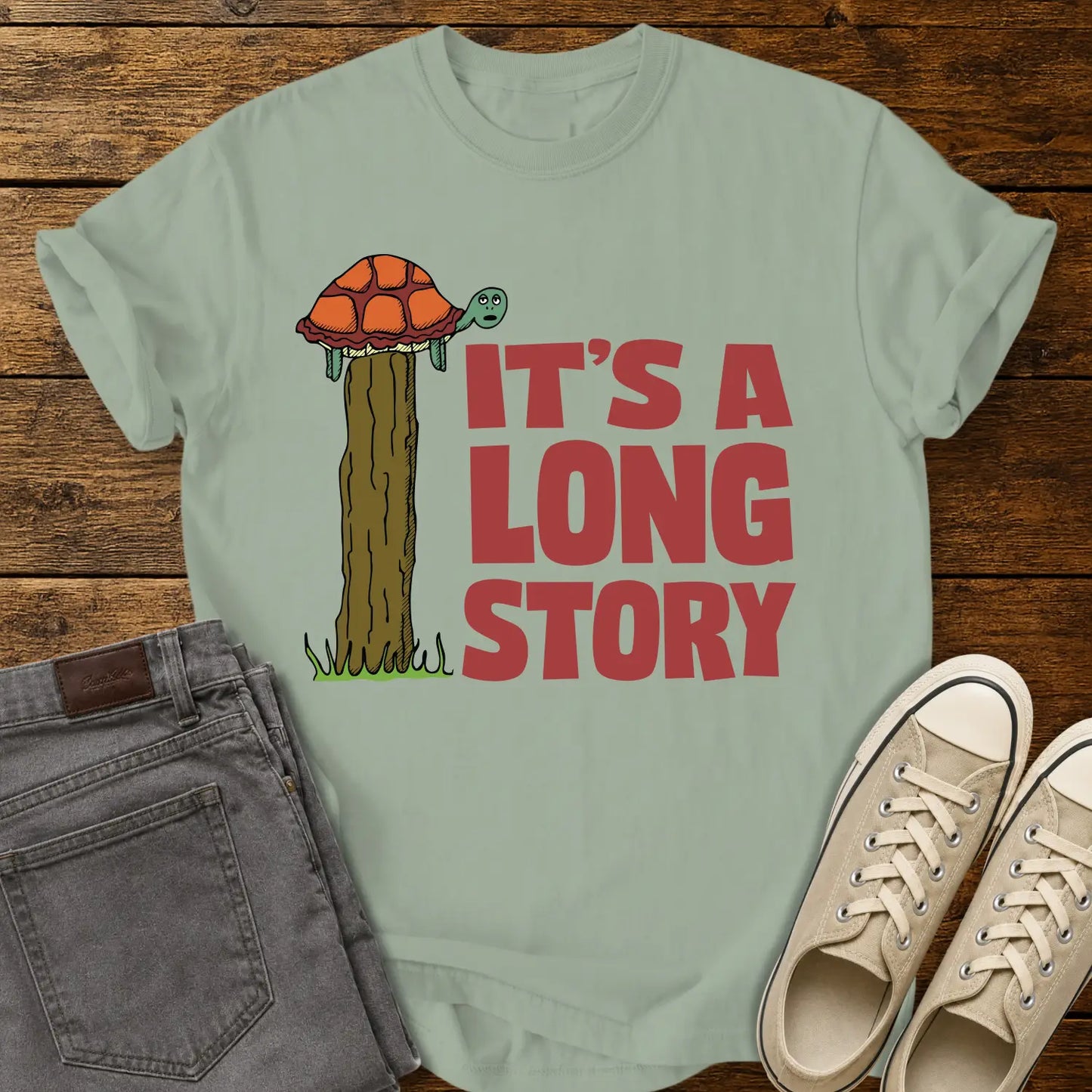 bay green T-shirt mockup featuring a humorous illustration of a turtle standing atop a fence post beside the phrase “IT’S A LONG STORY” in large vintage red lettering. The shirt is displayed on a rustic wood background alongside folded gray jeans and cream-colored canvas sneakers.