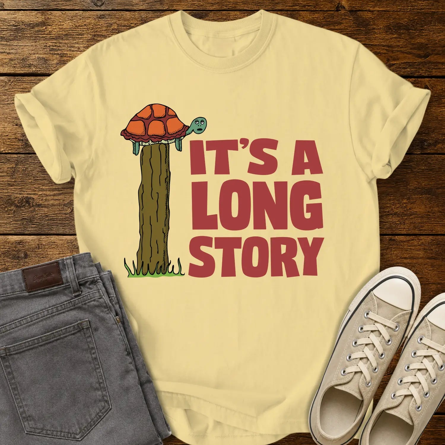 butter yellow T-shirt mockup featuring a humorous illustration of a turtle standing atop a fence post beside the phrase “IT’S A LONG STORY” in large vintage red lettering. The shirt is displayed on a rustic wood background alongside folded gray jeans and cream-colored canvas sneakers.