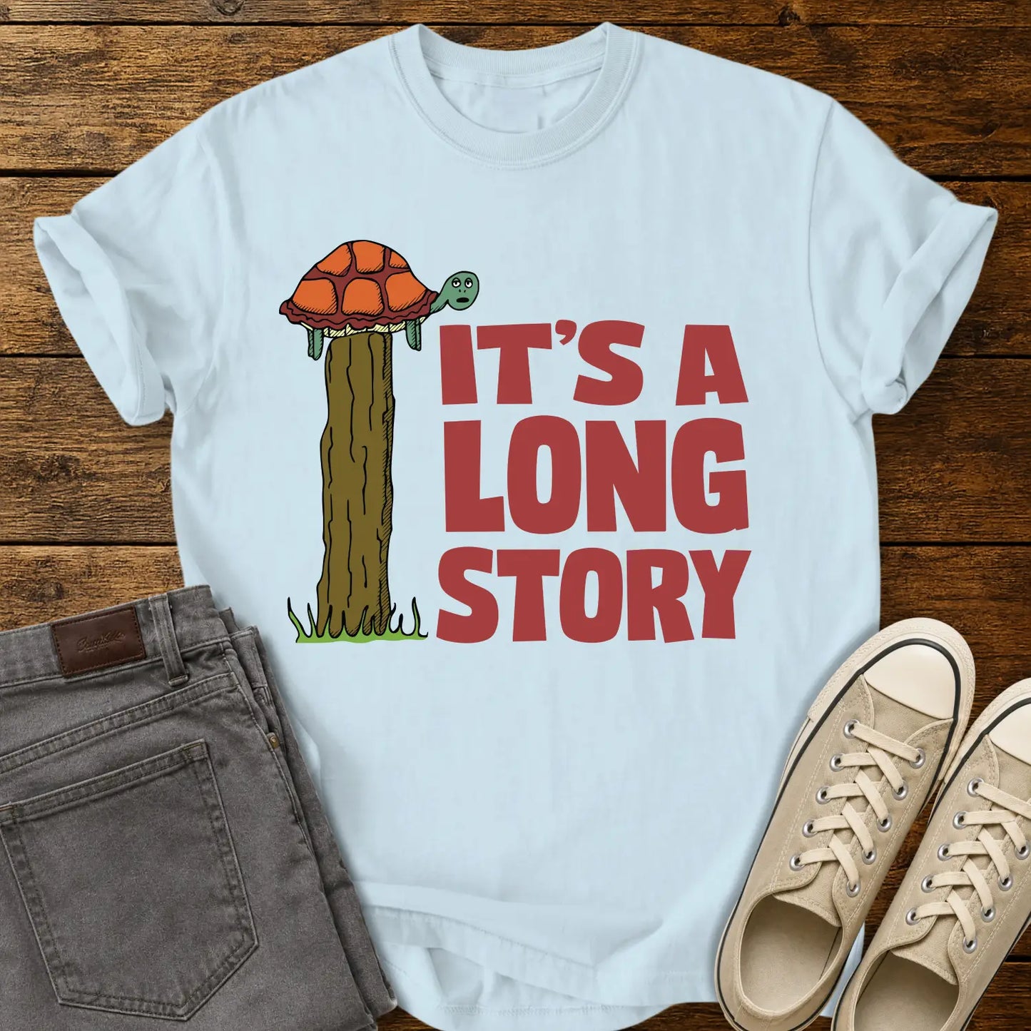 chambray blue T-shirt mockup featuring a humorous illustration of a turtle standing atop a fence post beside the phrase “IT’S A LONG STORY” in large vintage red lettering. The shirt is displayed on a rustic wood background alongside folded gray jeans and cream-colored canvas sneakers.