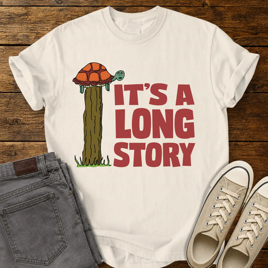 Ivory T-shirt mockup featuring a humorous illustration of a turtle standing atop a fence post beside the phrase “IT’S A LONG STORY” in large vintage red lettering. The shirt is displayed on a rustic wood background alongside folded gray jeans and cream-colored canvas sneakers.