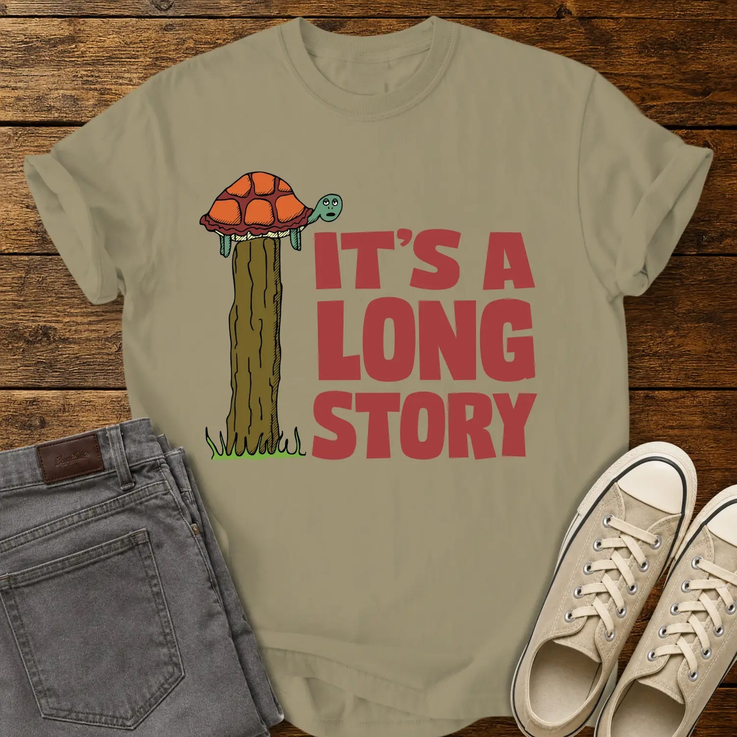 khaki T-shirt mockup featuring a humorous illustration of a turtle standing atop a fence post beside the phrase “IT’S A LONG STORY” in large vintage red lettering. The shirt is displayed on a rustic wood background alongside folded gray jeans and cream-colored canvas sneakers.