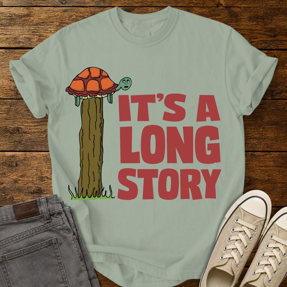 bay green graphic T-shirt mockup featuring a turtle sitting on top of a tall fence post beside the phrase “It’s a Long Story” in large red lettering, styled with gray jeans and beige sneakers on a rustic wood background.