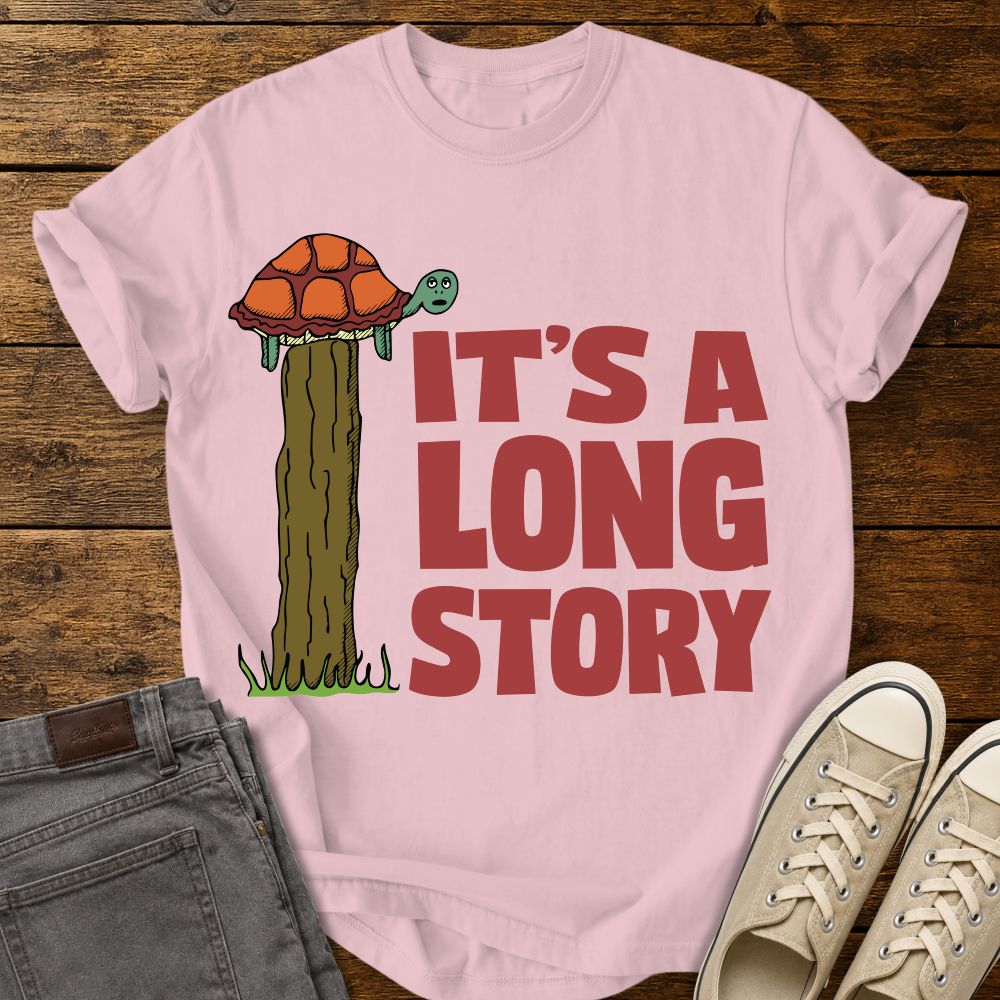blossom pink graphic T-shirt mockup featuring a turtle sitting on top of a tall fence post beside the phrase “It’s a Long Story” in large red lettering, styled with gray jeans and beige sneakers on a rustic wood background.