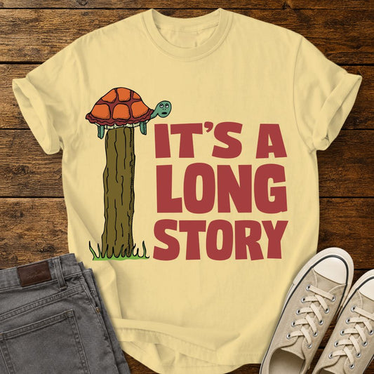 Butter yellow graphic T-shirt mockup featuring a turtle sitting on top of a tall fence post beside the phrase “It’s a Long Story” in large red lettering, styled with gray jeans and beige sneakers on a rustic wood background.