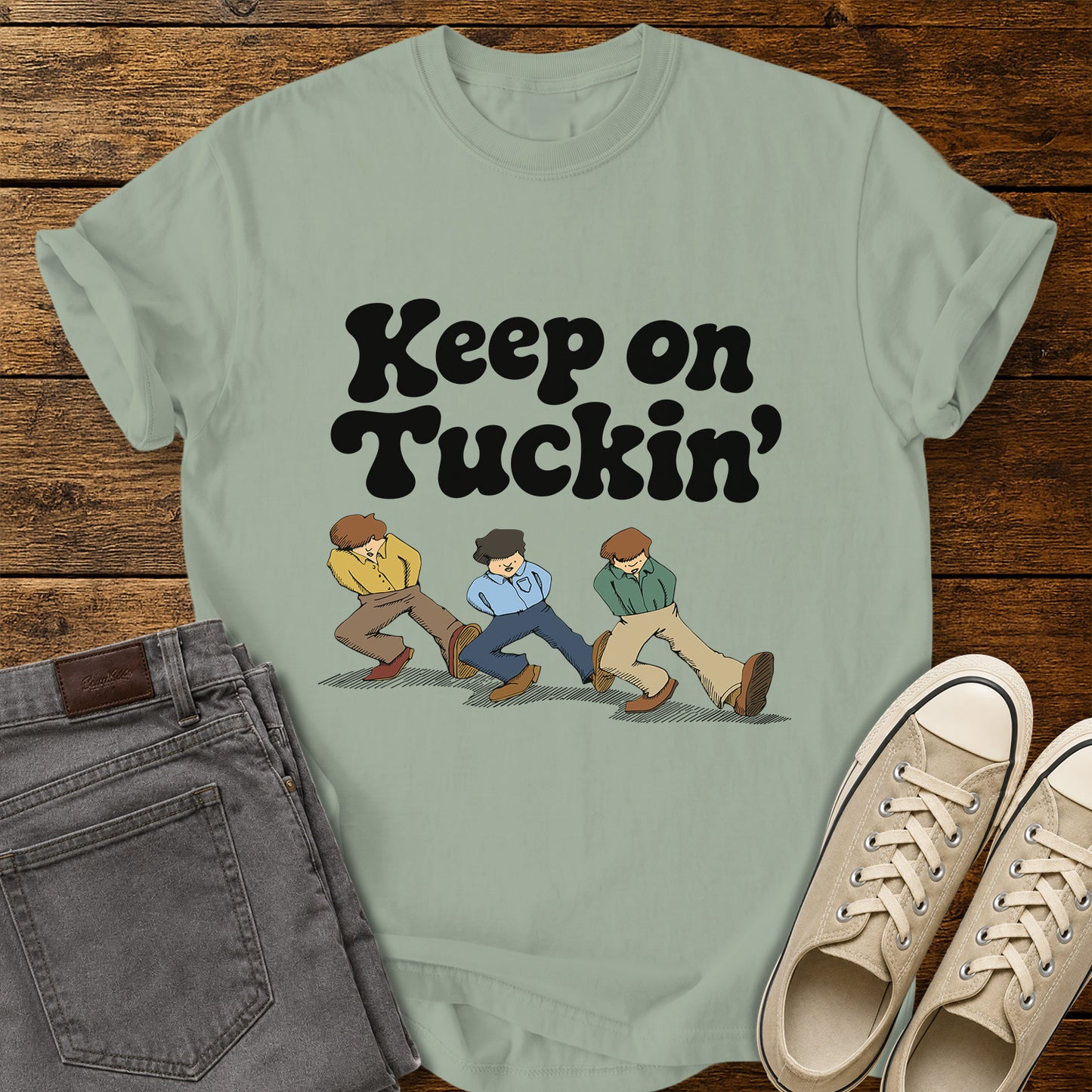 bay green flat-lay t-shirt mockup featuring bold retro black lettering that reads, "Keep on Tuckin'," above an illustration of three people tucking in their shirts while walking in a playful parody of the classic "Keep on Truckin'" pose. The vintage-inspired design is centered on the shirt, which is displayed on a rustic wood plank background alongside folded gray jeans and a pair of cream canvas sneakers.