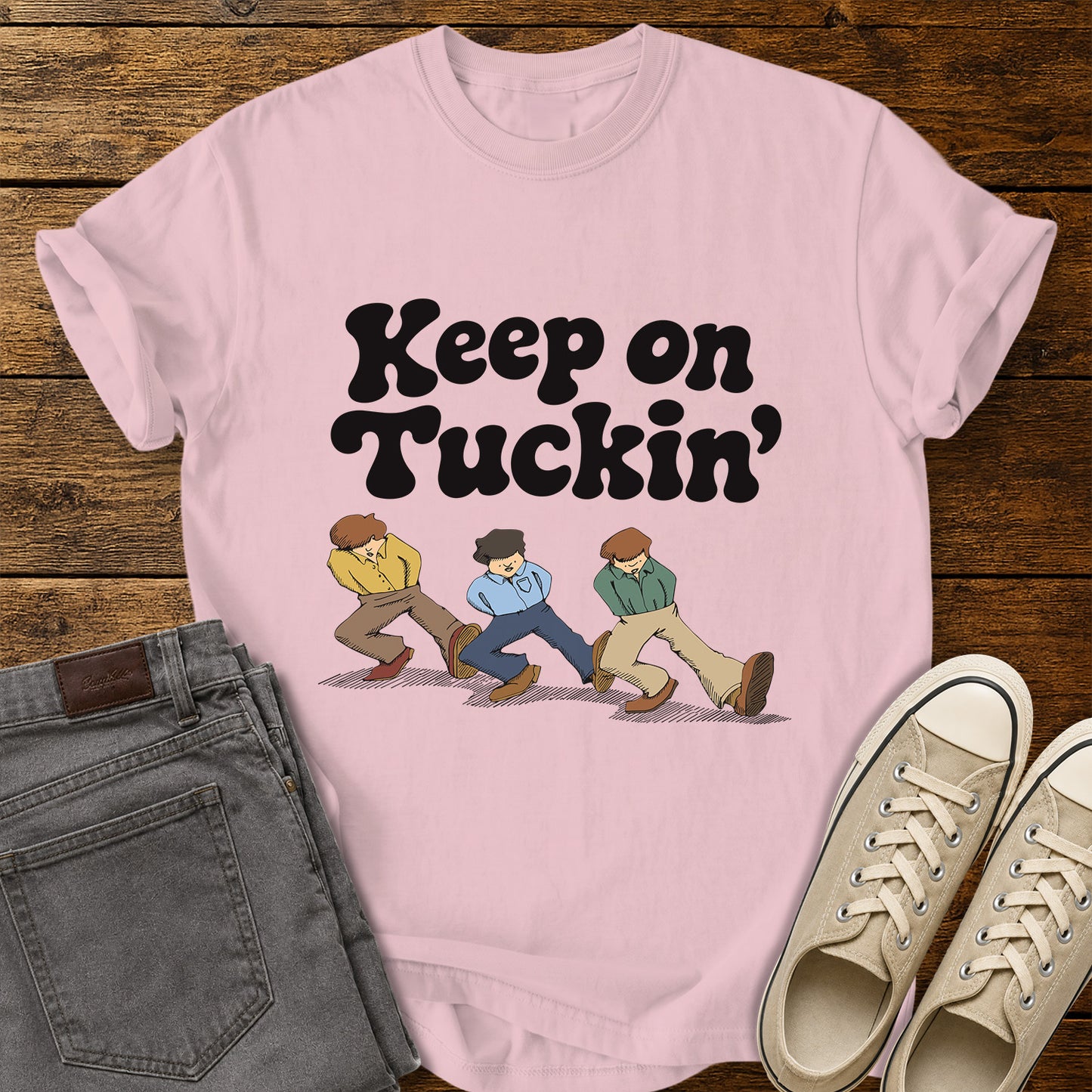 blossom pink flat-lay t-shirt mockup featuring bold retro black lettering that reads, "Keep on Tuckin'," above an illustration of three people tucking in their shirts while walking in a playful parody of the classic "Keep on Truckin'" pose. The vintage-inspired design is centered on the shirt, which is displayed on a rustic wood plank background alongside folded gray jeans and a pair of cream canvas sneakers.