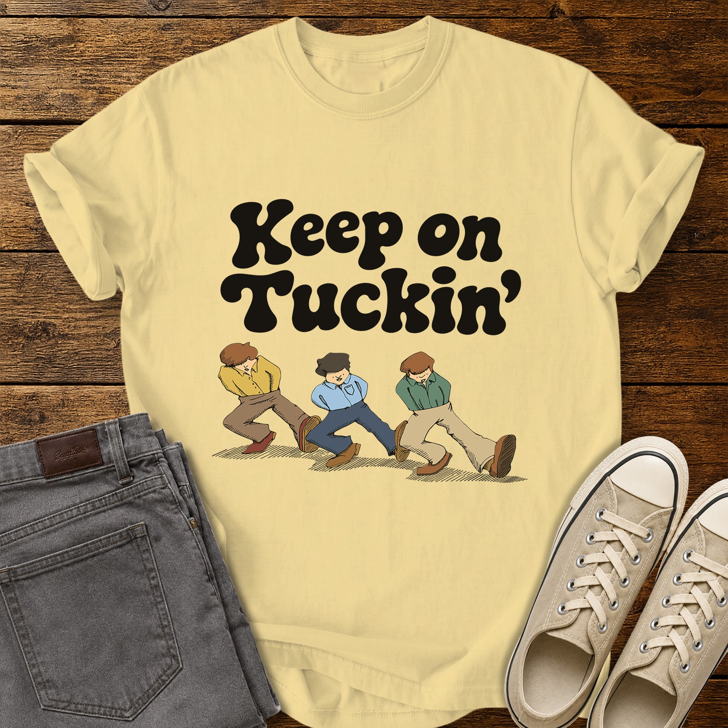 Butter yellow flat-lay t-shirt mockup featuring bold retro black lettering that reads, "Keep on Tuckin'," above an illustration of three people tucking in their shirts while walking in a playful parody of the classic "Keep on Truckin'" pose. The vintage-inspired design is centered on the shirt, which is displayed on a rustic wood plank background alongside folded gray jeans and a pair of cream canvas sneakers.