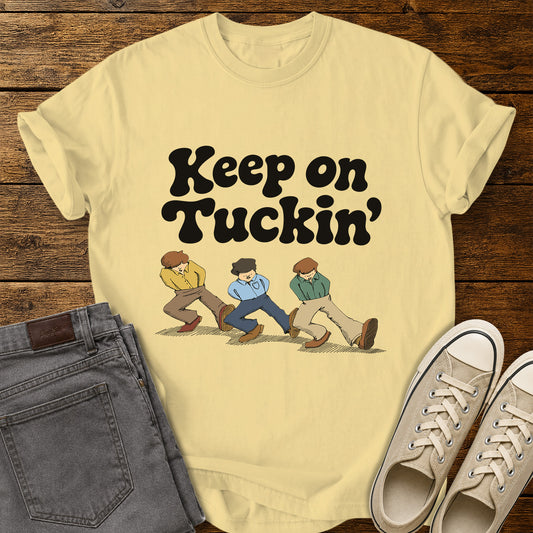 Butter yellow flat-lay t-shirt mockup featuring bold retro black lettering that reads, "Keep on Tuckin'," above an illustration of three people tucking in their shirts while walking in a playful parody of the classic "Keep on Truckin'" pose. The vintage-inspired design is centered on the shirt, which is displayed on a rustic wood plank background alongside folded gray jeans and a pair of cream canvas sneakers.