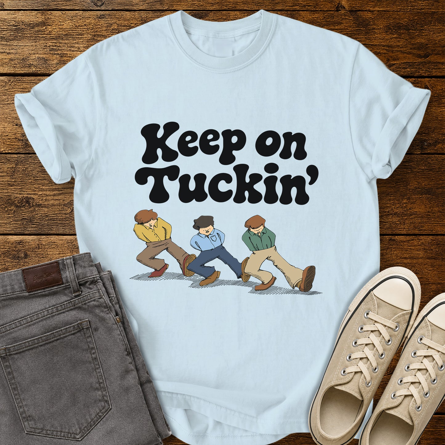 chambray blue flat-lay t-shirt mockup featuring bold retro black lettering that reads, "Keep on Tuckin'," above an illustration of three people tucking in their shirts while walking in a playful parody of the classic "Keep on Truckin'" pose. The vintage-inspired design is centered on the shirt, which is displayed on a rustic wood plank background alongside folded gray jeans and a pair of cream canvas sneakers.