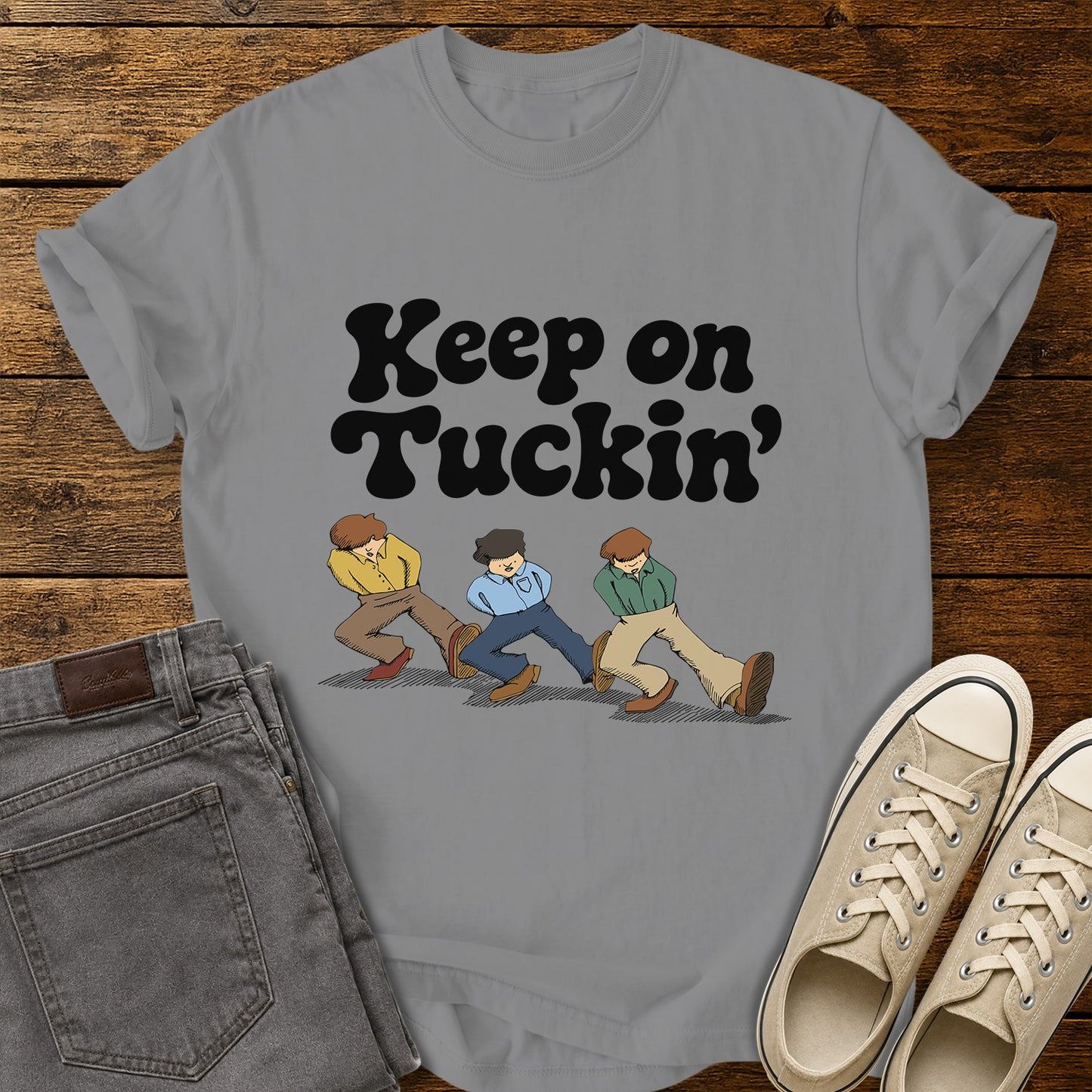 grey flat-lay t-shirt mockup featuring bold retro black lettering that reads, "Keep on Tuckin'," above an illustration of three people tucking in their shirts while walking in a playful parody of the classic "Keep on Truckin'" pose. The vintage-inspired design is centered on the shirt, which is displayed on a rustic wood plank background alongside folded gray jeans and a pair of cream canvas sneakers.