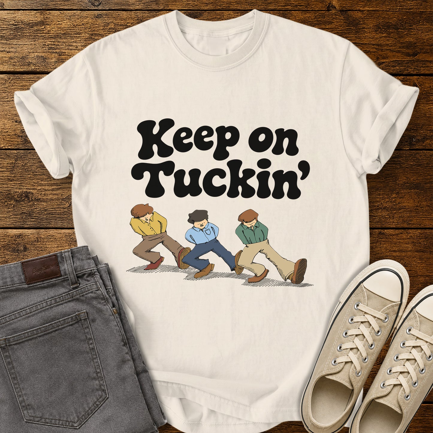 ivory flat-lay t-shirt mockup featuring bold retro black lettering that reads, "Keep on Tuckin'," above an illustration of three people tucking in their shirts while walking in a playful parody of the classic "Keep on Truckin'" pose. The vintage-inspired design is centered on the shirt, which is displayed on a rustic wood plank background alongside folded gray jeans and a pair of cream canvas sneakers.