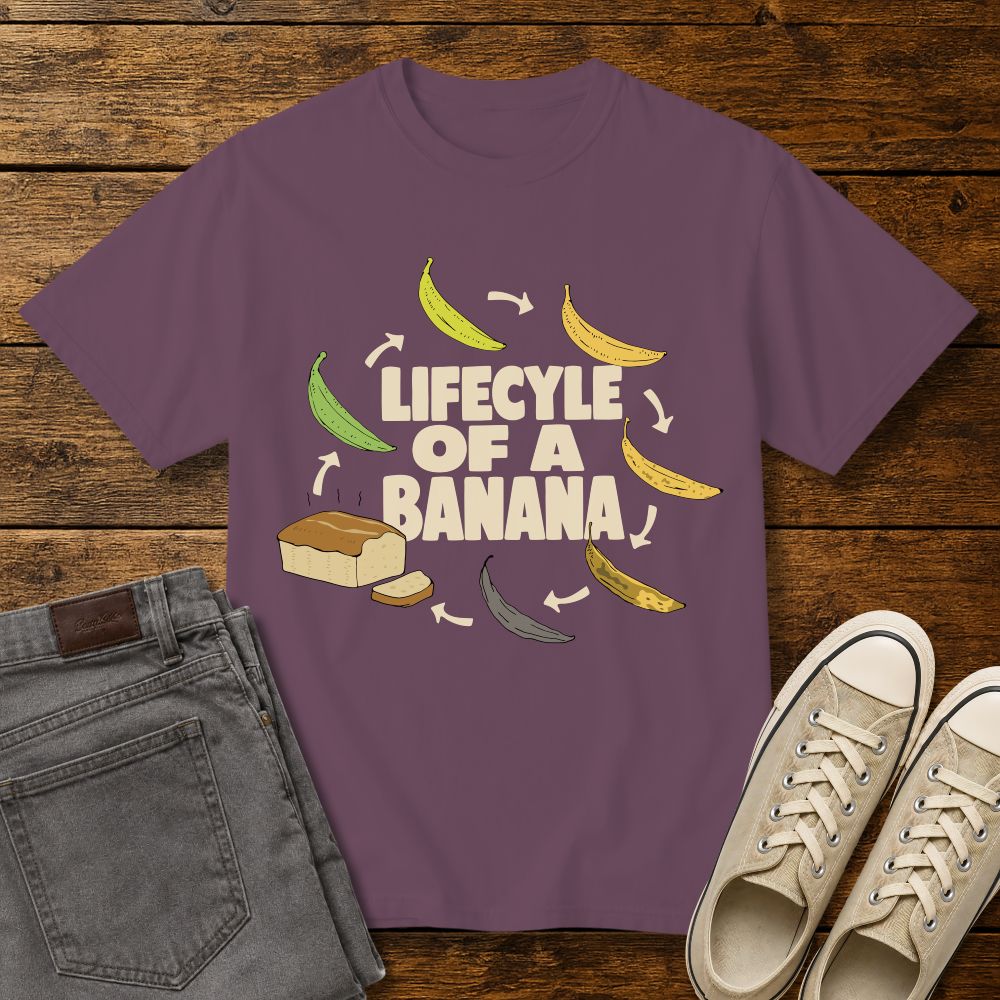 berry color graphic T-shirt mockup featuring the phrase “Lifecycle of a Banana” surrounded by illustrations of bananas ripening into banana bread, styled with gray jeans and beige sneakers on a rustic wood background.