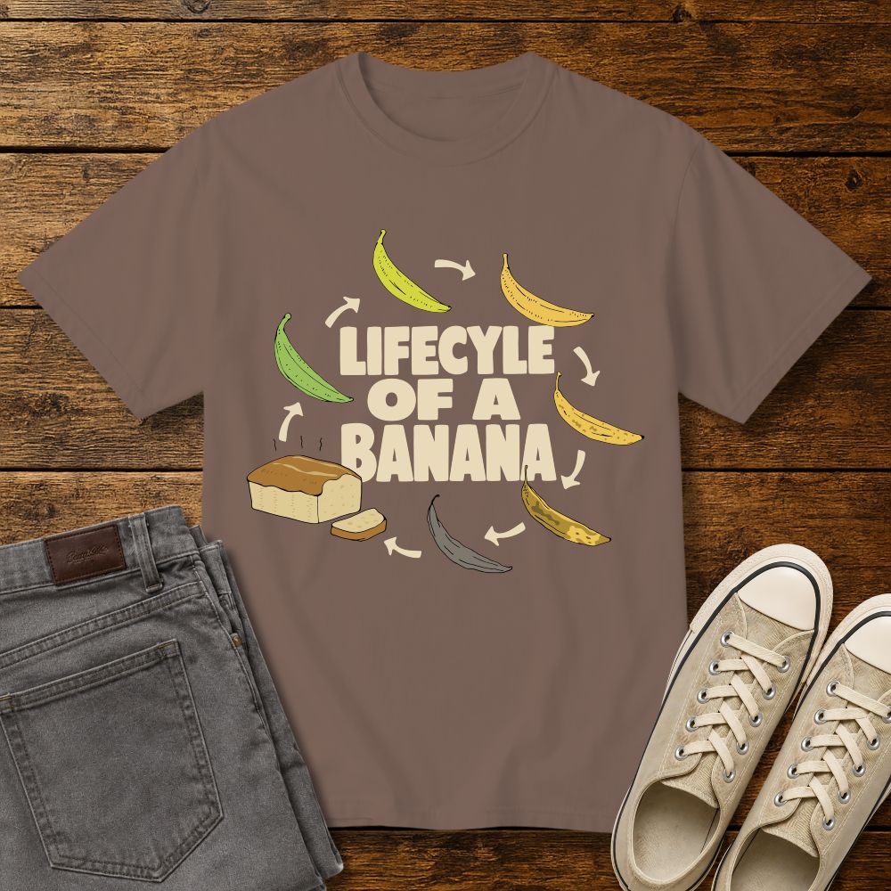 espresso brown graphic T-shirt mockup featuring the phrase “Lifecycle of a Banana” surrounded by illustrations of bananas ripening into banana bread, styled with gray jeans and beige sneakers on a rustic wood background.