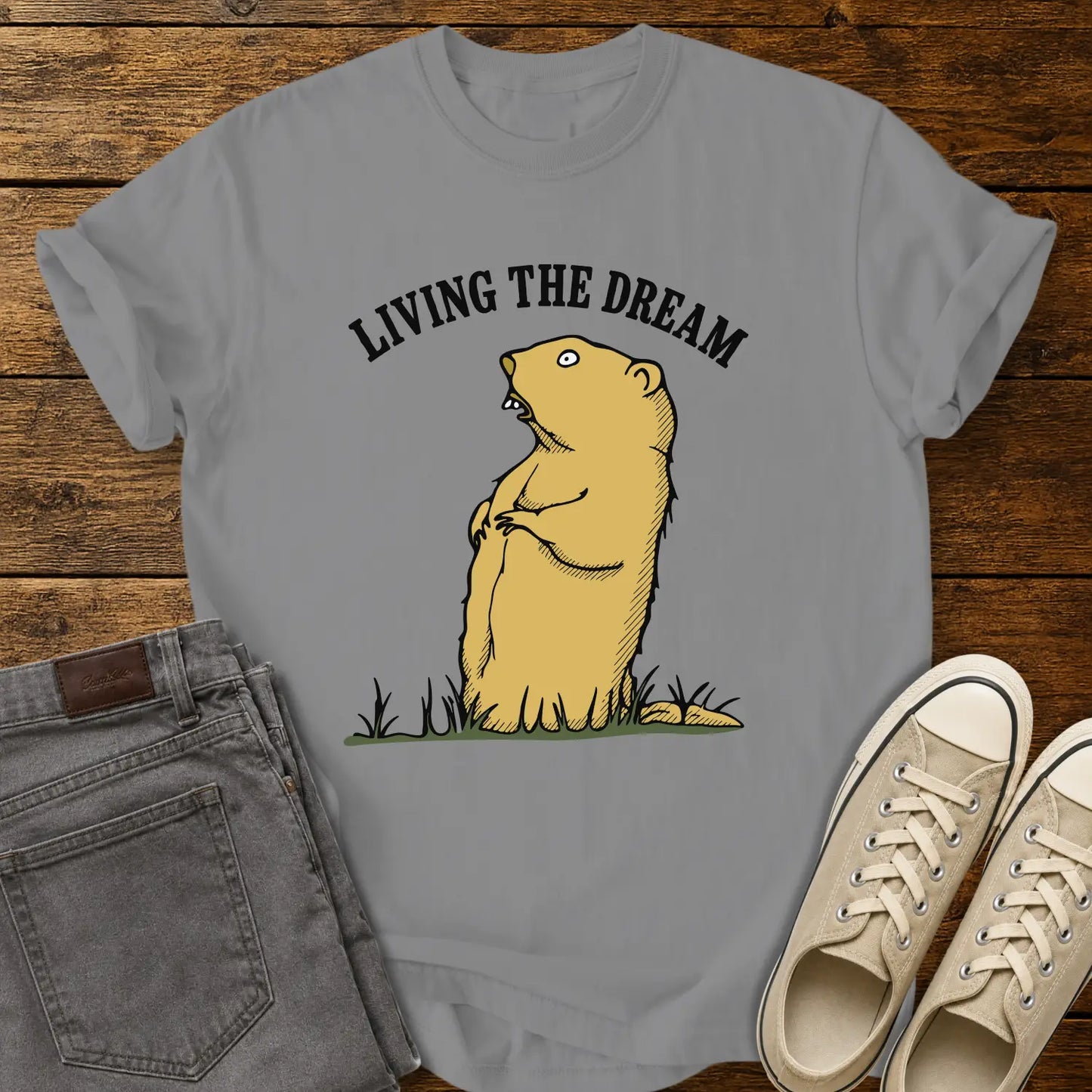 grey graphic T-shirt mockup featuring a standing groundhog illustration beneath the phrase “Living the Dream” in bold black arched lettering, styled with gray jeans and beige sneakers on a rustic wood background.