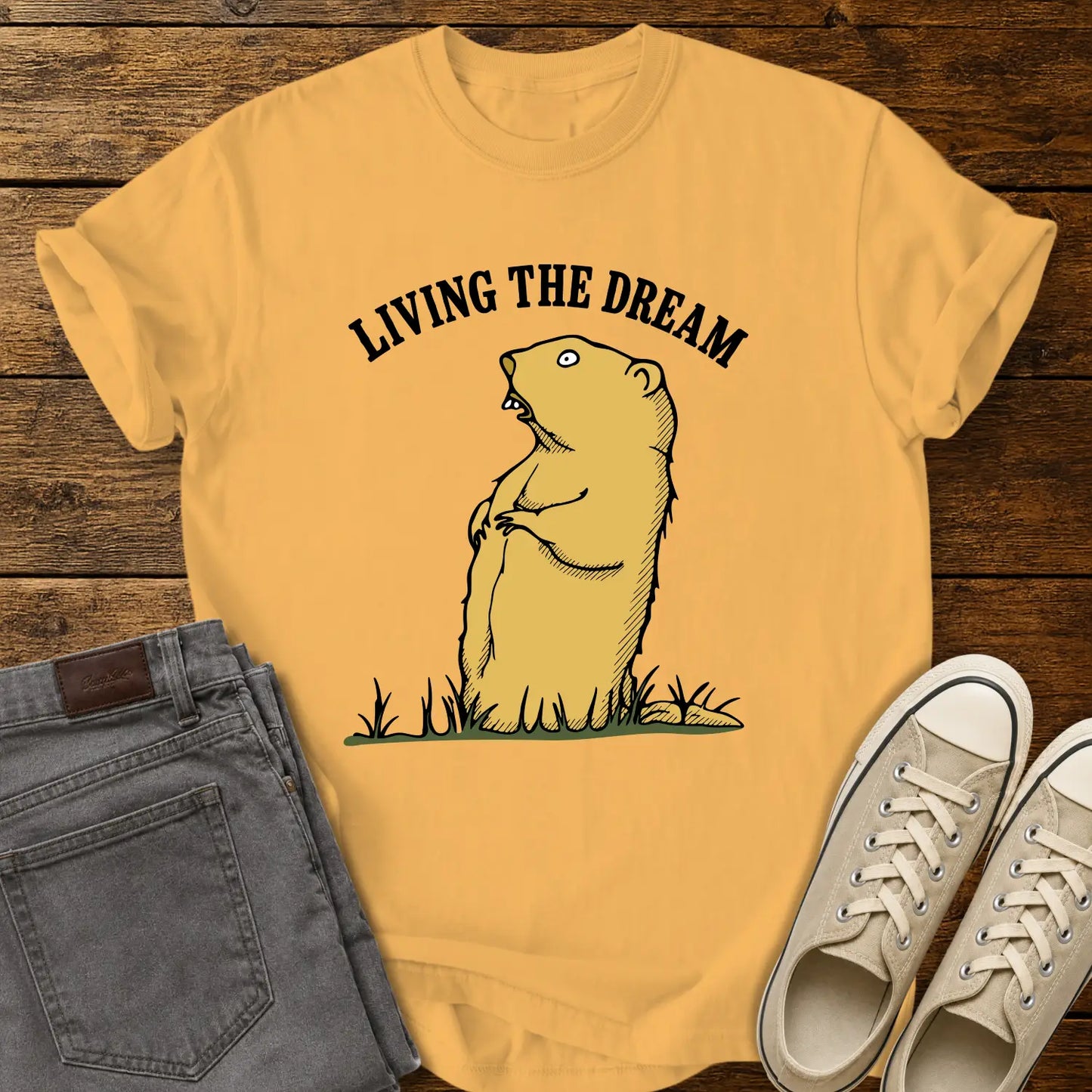 citrus yellow graphic T-shirt mockup featuring a standing groundhog illustration beneath the phrase “Living the Dream” in bold black arched lettering, styled with gray jeans and beige sneakers on a rustic wood background.