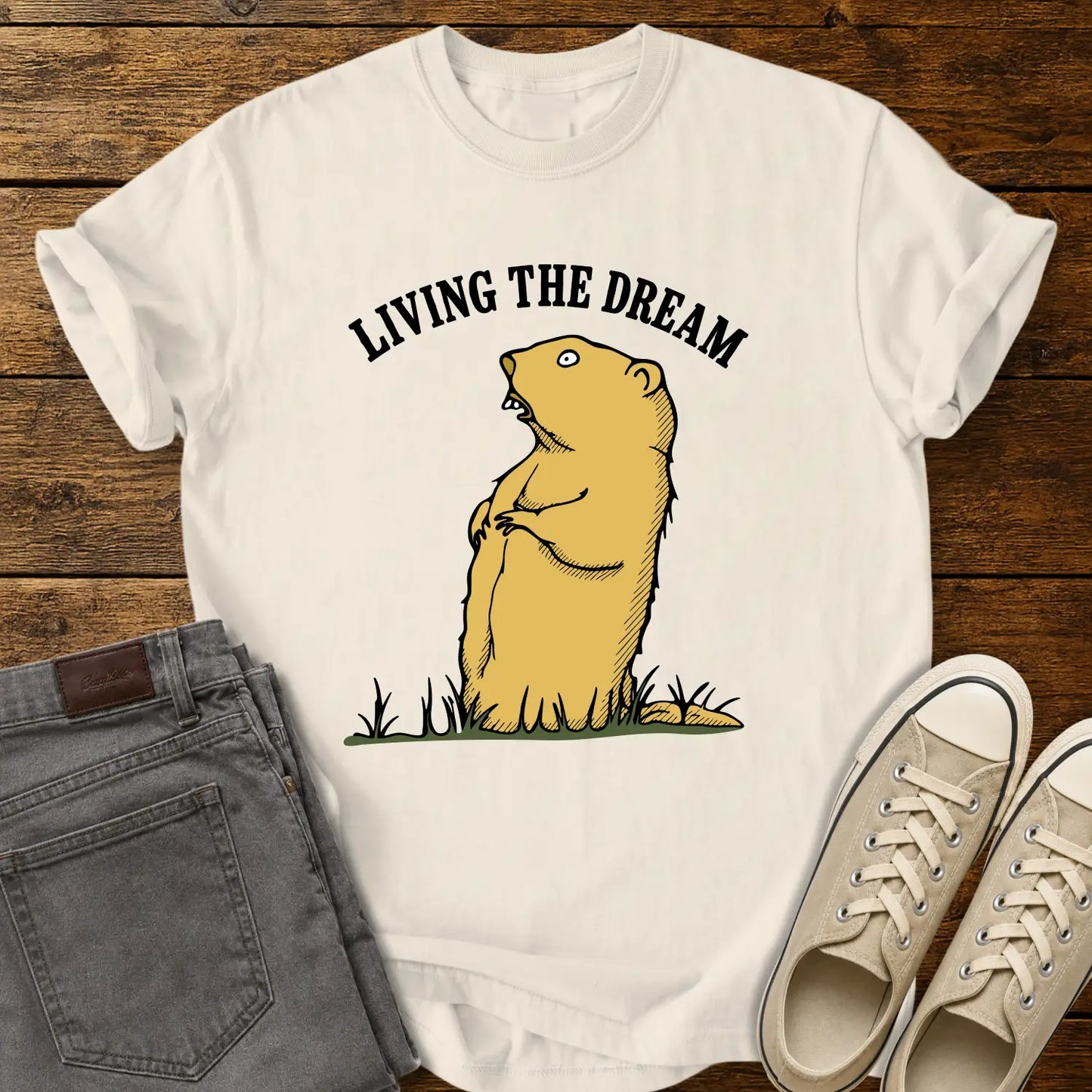 ivory graphic T-shirt mockup featuring a standing groundhog illustration beneath the phrase “Living the Dream” in bold black arched lettering, styled with gray jeans and beige sneakers on a rustic wood background.