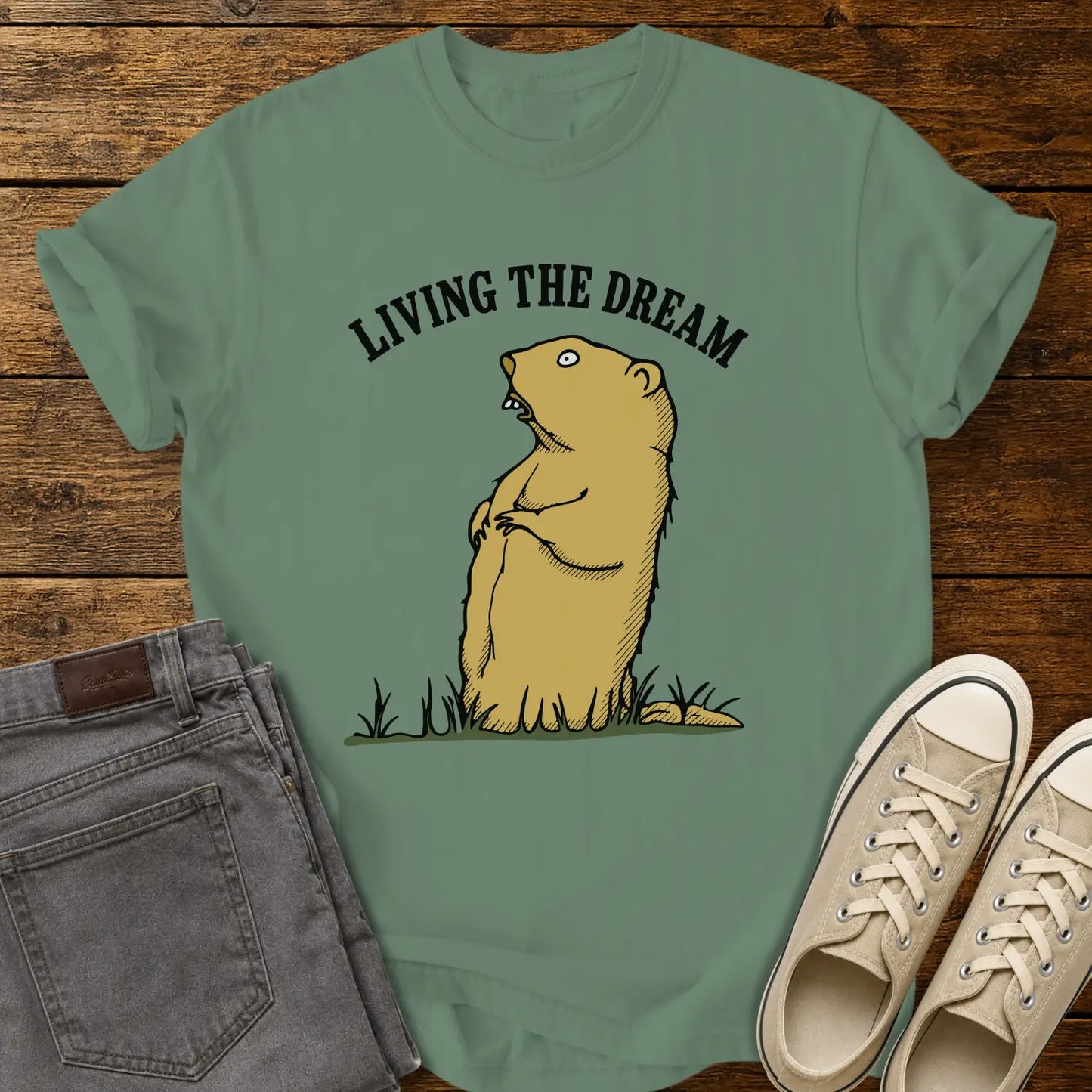light green graphic T-shirt mockup featuring a standing groundhog illustration beneath the phrase “Living the Dream” in bold black arched lettering, styled with gray jeans and beige sneakers on a rustic wood background.