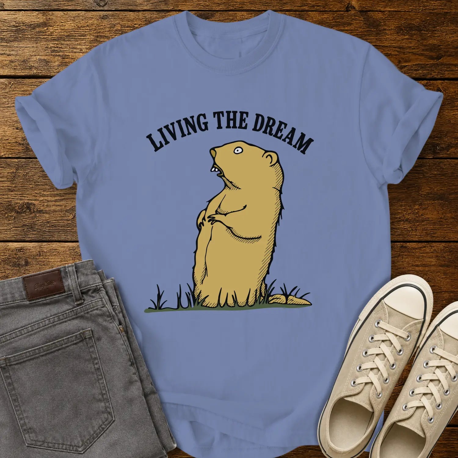 Washed denim blue graphic T-shirt mockup featuring a standing groundhog illustration beneath the phrase “Living the Dream” in bold black arched lettering, styled with gray jeans and beige sneakers on a rustic wood background.