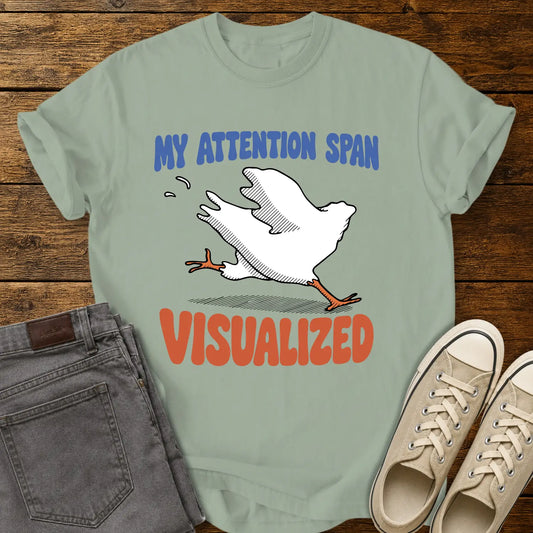 Bay green T-shirt featuring the humorous phrase “MY ATTENTION SPAN VISUALIZED” with a cartoon headless chicken running across the center. The design uses blue and orange retro lettering with a playful illustration that conveys distracted or short attention span humor. The shirt is displayed on a rustic wood plank background alongside folded gray jeans and cream canvas sneakers.