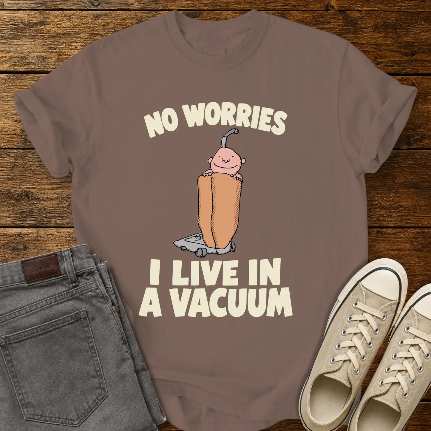 espresso brown graphic T-shirt mockup featuring a cartoon person riding inside a vacuum cleaner bag with the phrase “No Worries, I Live in a Vacuum” in bold white lettering, styled with gray jeans and beige sneakers on a rustic wood background.