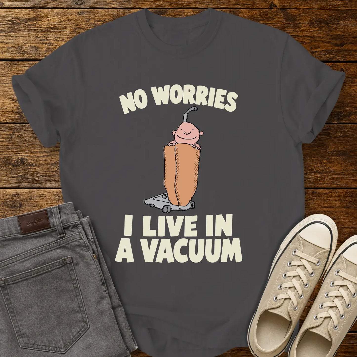 graphite grey graphic T-shirt mockup featuring a cartoon person riding inside a vacuum cleaner bag with the phrase “No Worries, I Live in a Vacuum” in bold white lettering, styled with gray jeans and beige sneakers on a rustic wood background.