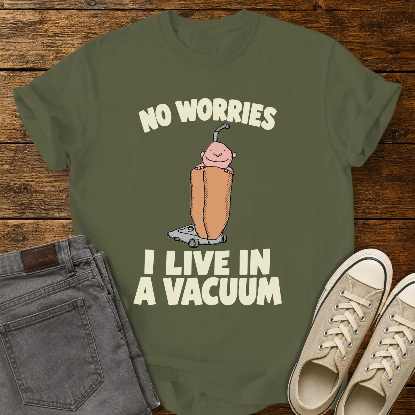 hemp green graphic T-shirt mockup featuring a cartoon person riding inside a vacuum cleaner bag with the phrase “No Worries, I Live in a Vacuum” in bold white lettering, styled with gray jeans and beige sneakers on a rustic wood background.