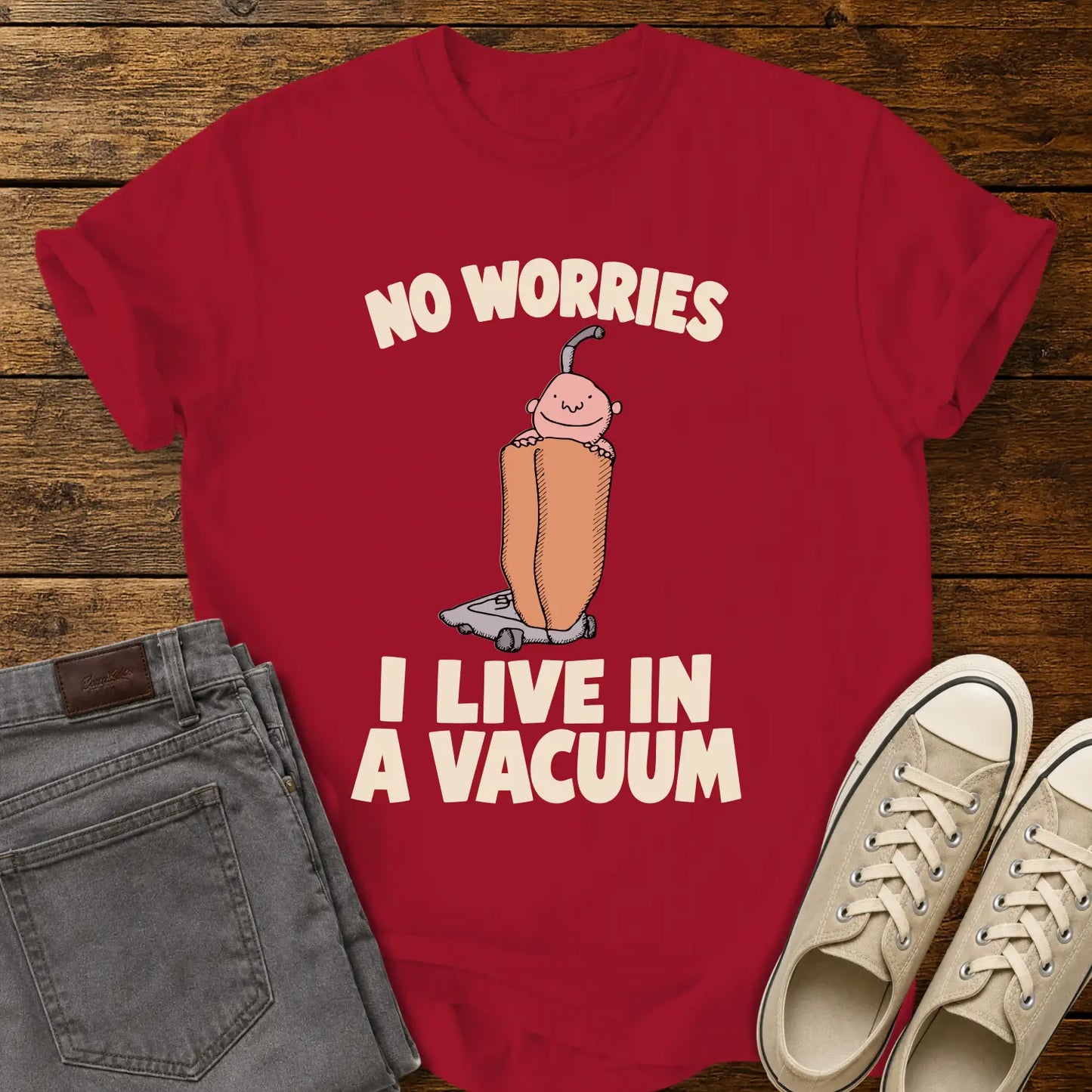 red graphic T-shirt mockup featuring a cartoon person riding inside a vacuum cleaner bag with the phrase “No Worries, I Live in a Vacuum” in bold white lettering, styled with gray jeans and beige sneakers on a rustic wood background.