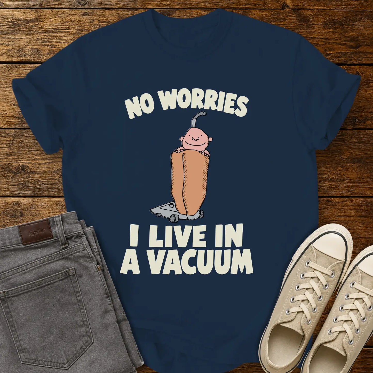 True navy graphic T-shirt mockup featuring a cartoon person riding inside a vacuum cleaner bag with the phrase “No Worries, I Live in a Vacuum” in bold white lettering, styled with gray jeans and beige sneakers on a rustic wood background.
