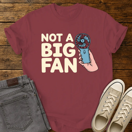 Chili red T-shirt mockup featuring the phrase “NOT A BIG FAN” in large cream-colored lettering beside an illustration of a hand holding a small blue portable fan. The shirt is displayed on a rustic wood background alongside folded gray jeans and cream-colored canvas sneakers.
