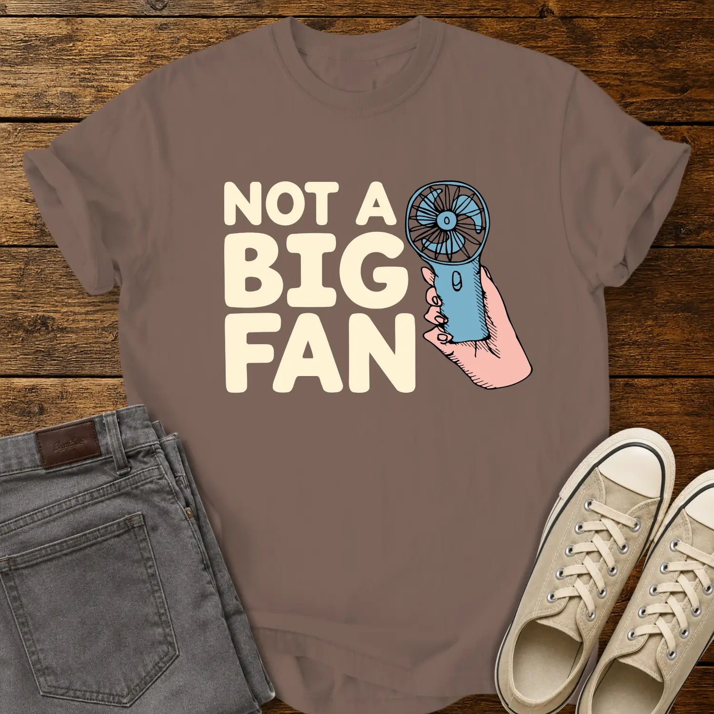 espresso brown T-shirt mockup featuring the phrase “NOT A BIG FAN” in large cream-colored lettering beside an illustration of a hand holding a small blue portable fan. The shirt is displayed on a rustic wood background alongside folded gray jeans and cream-colored canvas sneakers.