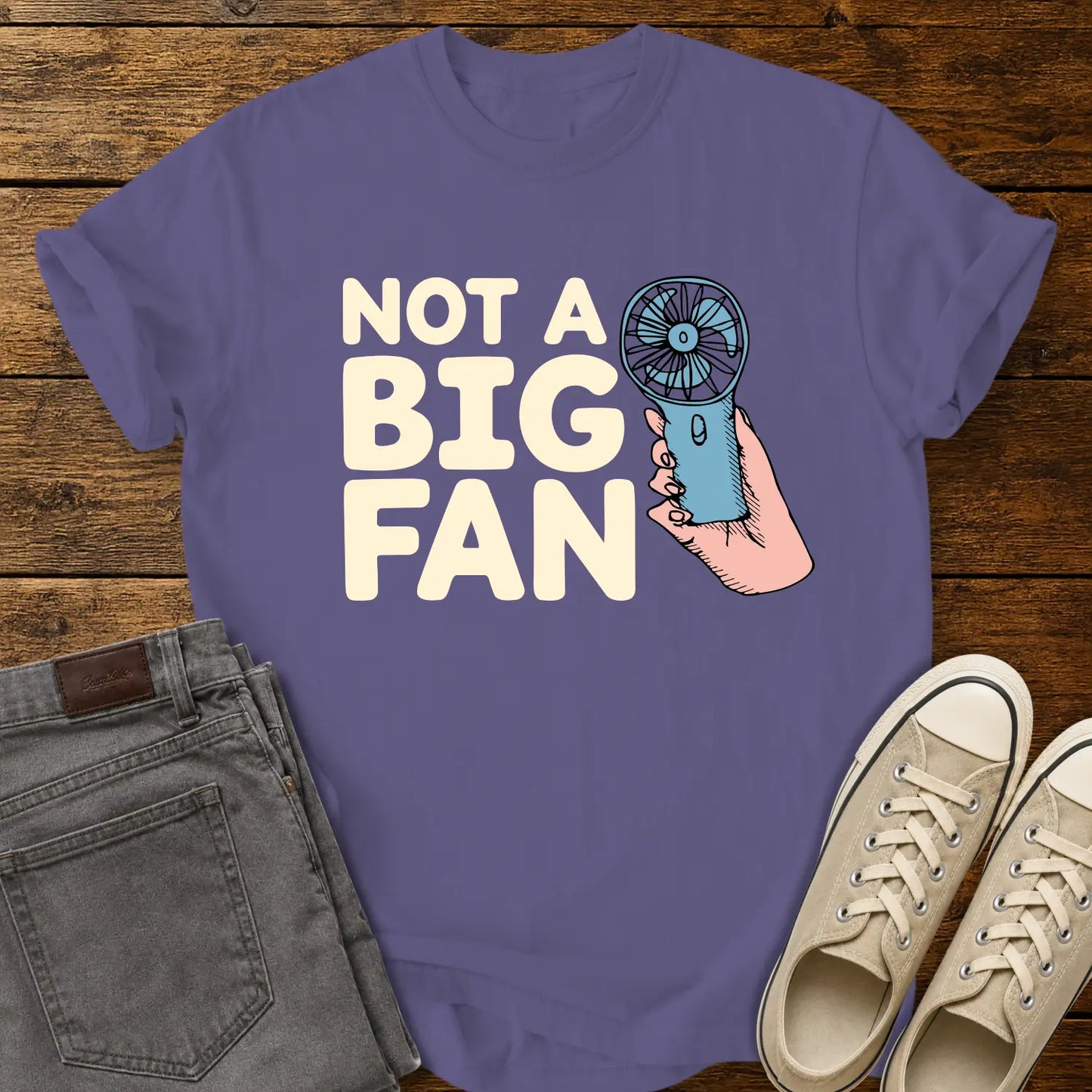 grape T-shirt mockup featuring the phrase “NOT A BIG FAN” in large cream-colored lettering beside an illustration of a hand holding a small blue portable fan. The shirt is displayed on a rustic wood background alongside folded gray jeans and cream-colored canvas sneakers.