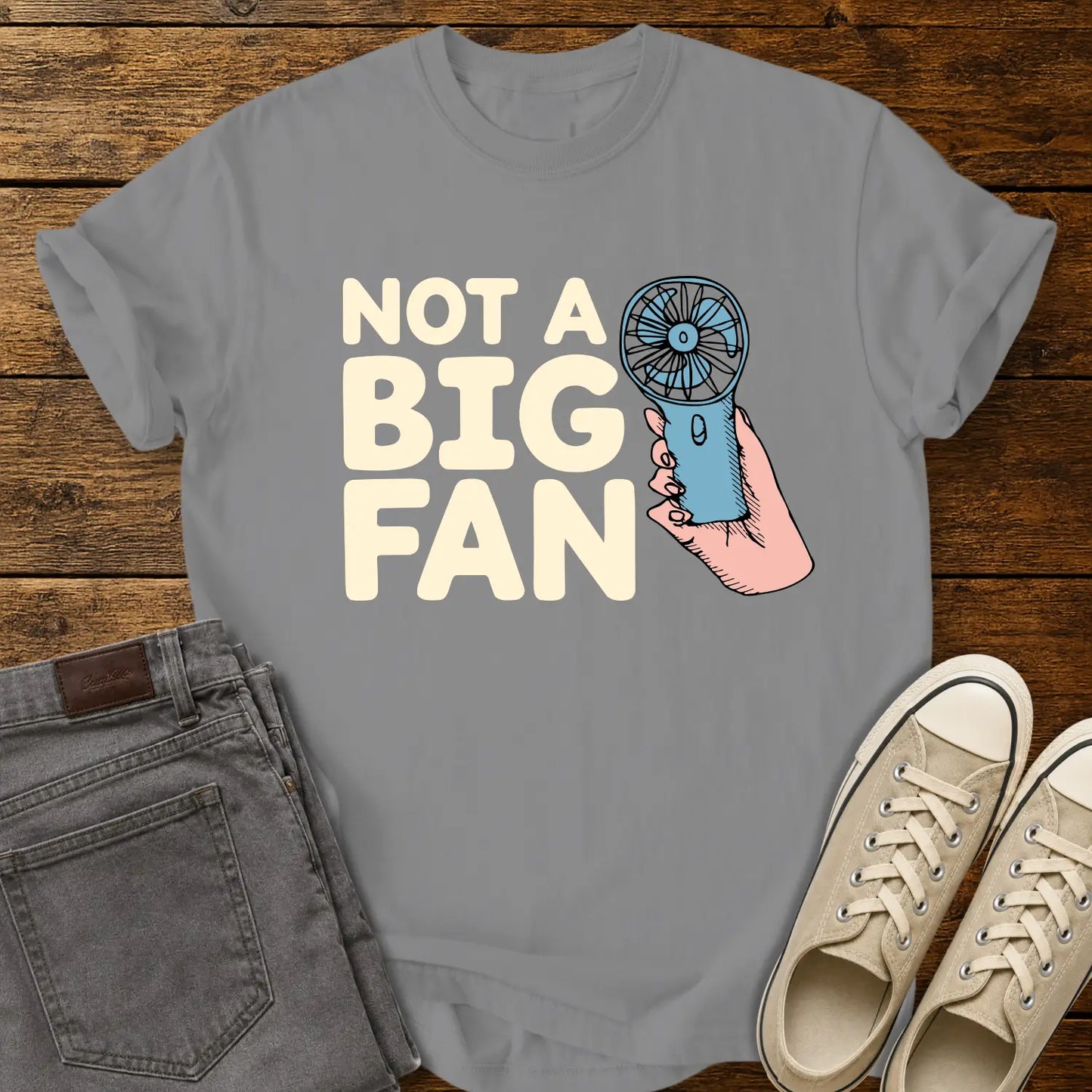 grey T-shirt mockup featuring the phrase “NOT A BIG FAN” in large cream-colored lettering beside an illustration of a hand holding a small blue portable fan. The shirt is displayed on a rustic wood background alongside folded gray jeans and cream-colored canvas sneakers.