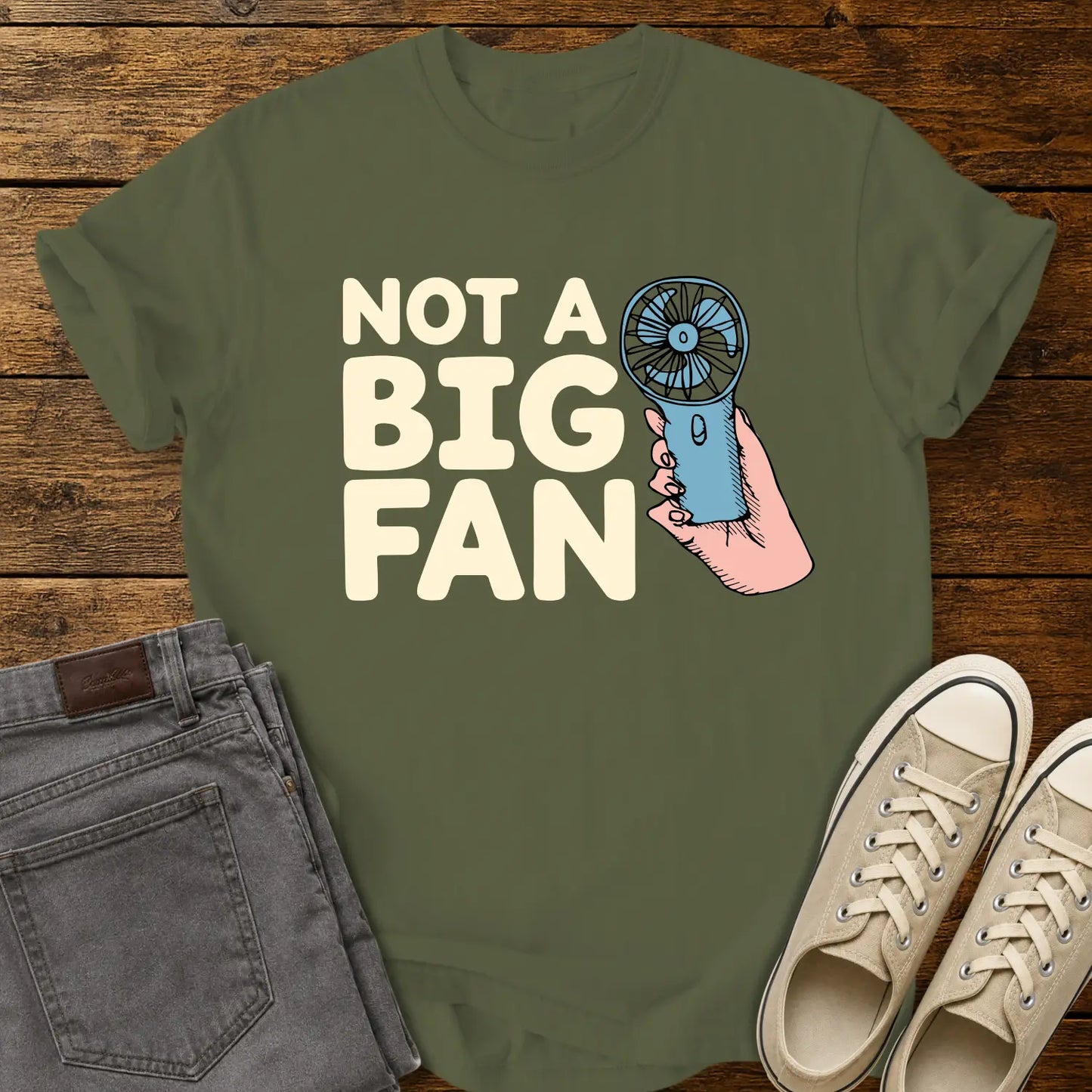 hemp green T-shirt mockup featuring the phrase “NOT A BIG FAN” in large cream-colored lettering beside an illustration of a hand holding a small blue portable fan. The shirt is displayed on a rustic wood background alongside folded gray jeans and cream-colored canvas sneakers.