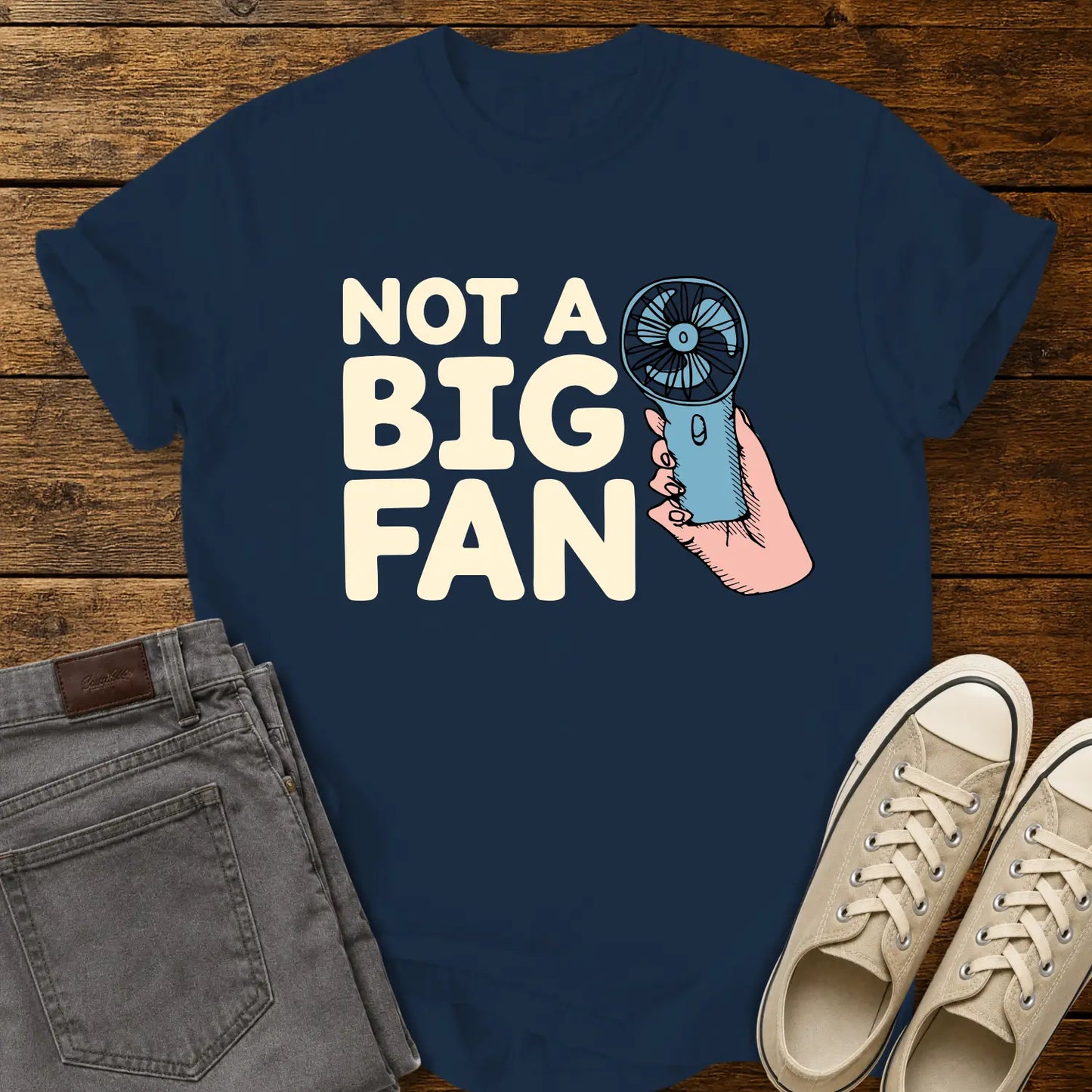 true navy blue T-shirt mockup featuring the phrase “NOT A BIG FAN” in large cream-colored lettering beside an illustration of a hand holding a small blue portable fan. The shirt is displayed on a rustic wood background alongside folded gray jeans and cream-colored canvas sneakers.