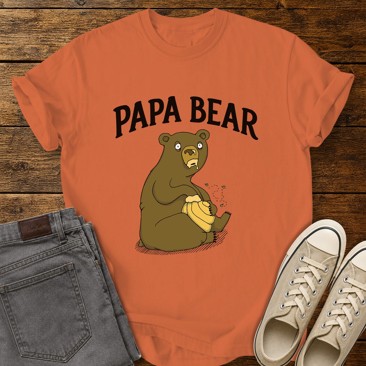 burnt orange flat-lay t-shirt mockup featuring a humorous cartoon bear sitting with a honey pot while bees buzz around it. Bold black lettering above the illustration reads, "PAPA BEAR." The vintage-inspired design is centered on the shirt, which is displayed on a rustic wood plank background alongside folded gray jeans and a pair of cream canvas sneakers.