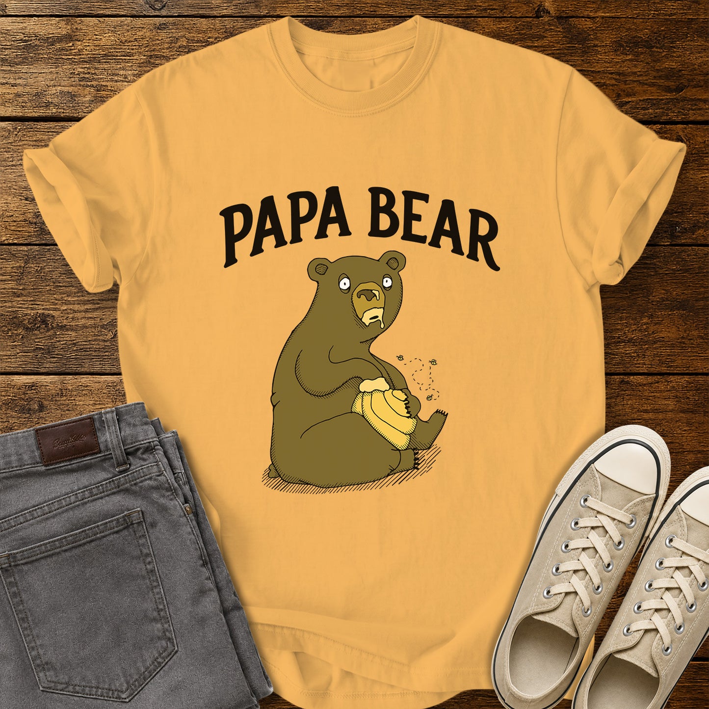 citrus yellow flat-lay t-shirt mockup featuring a humorous cartoon bear sitting with a honey pot while bees buzz around it. Bold black lettering above the illustration reads, "PAPA BEAR." The vintage-inspired design is centered on the shirt, which is displayed on a rustic wood plank background alongside folded gray jeans and a pair of cream canvas sneakers.