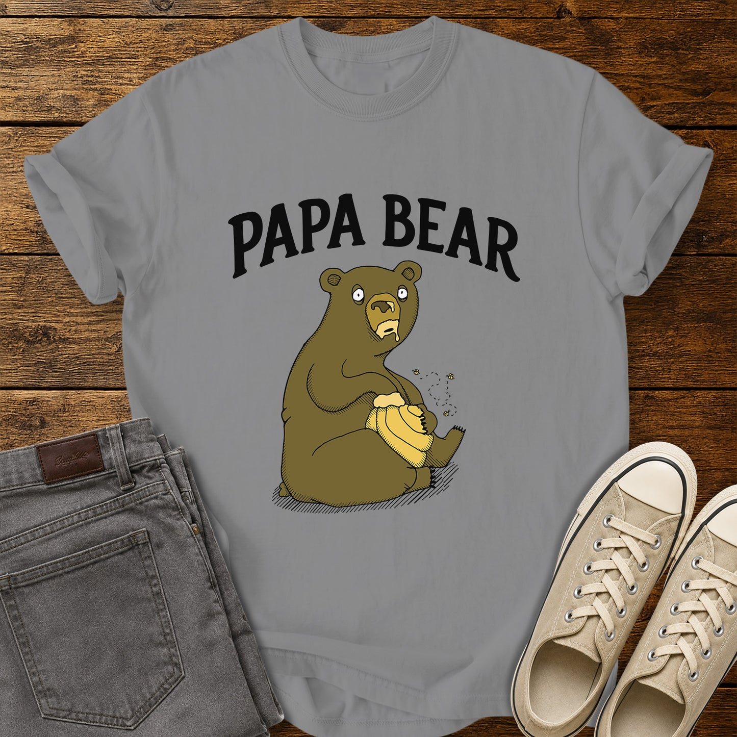 grey flat-lay t-shirt mockup featuring a humorous cartoon bear sitting with a honey pot while bees buzz around it. Bold black lettering above the illustration reads, "PAPA BEAR." The vintage-inspired design is centered on the shirt, which is displayed on a rustic wood plank background alongside folded gray jeans and a pair of cream canvas sneakers.