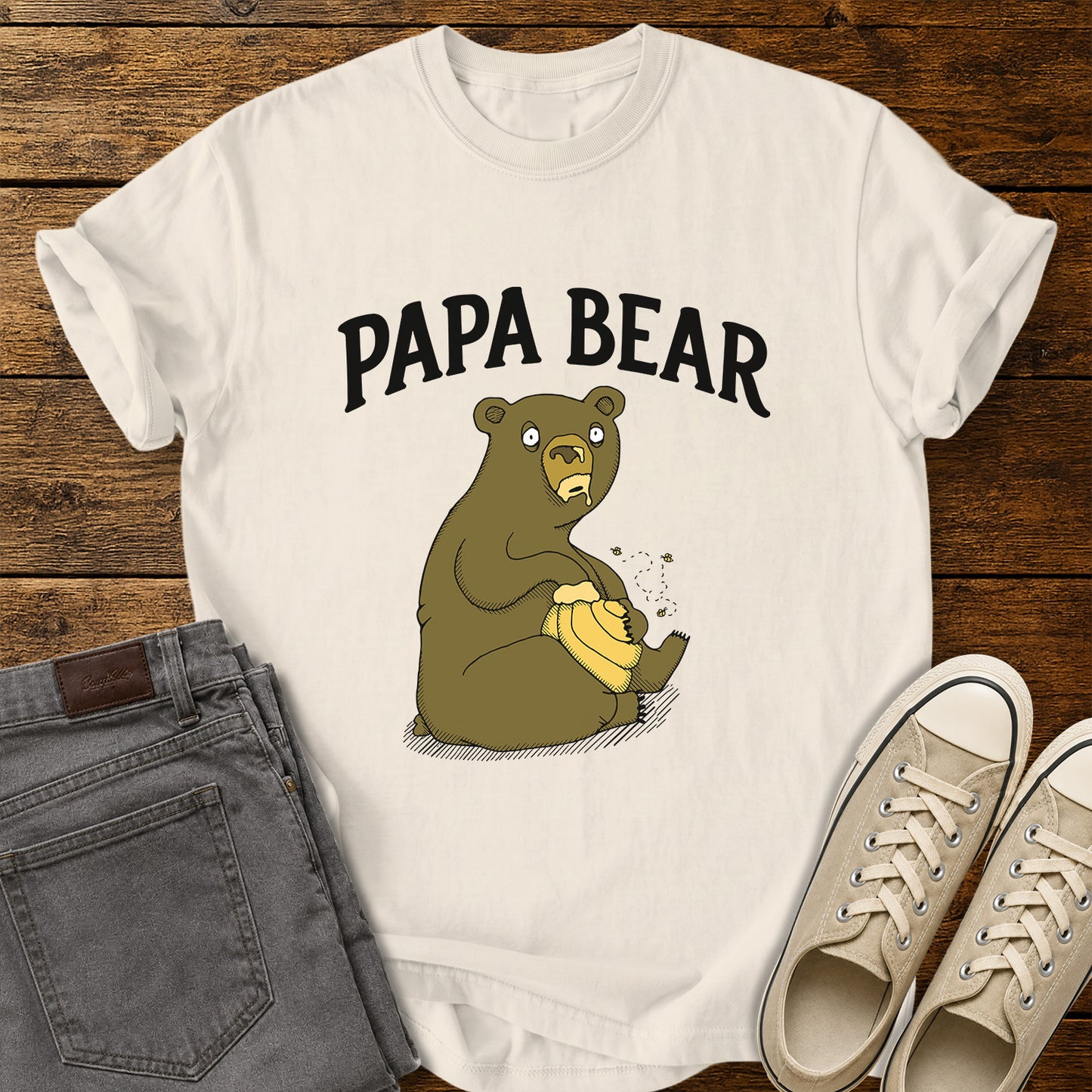 ivory flat-lay t-shirt mockup featuring a humorous cartoon bear sitting with a honey pot while bees buzz around it. Bold black lettering above the illustration reads, "PAPA BEAR." The vintage-inspired design is centered on the shirt, which is displayed on a rustic wood plank background alongside folded gray jeans and a pair of cream canvas sneakers.