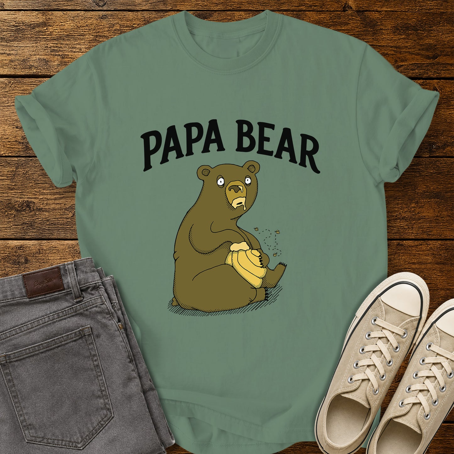 light green flat-lay t-shirt mockup featuring a humorous cartoon bear sitting with a honey pot while bees buzz around it. Bold black lettering above the illustration reads, "PAPA BEAR." The vintage-inspired design is centered on the shirt, which is displayed on a rustic wood plank background alongside folded gray jeans and a pair of cream canvas sneakers.