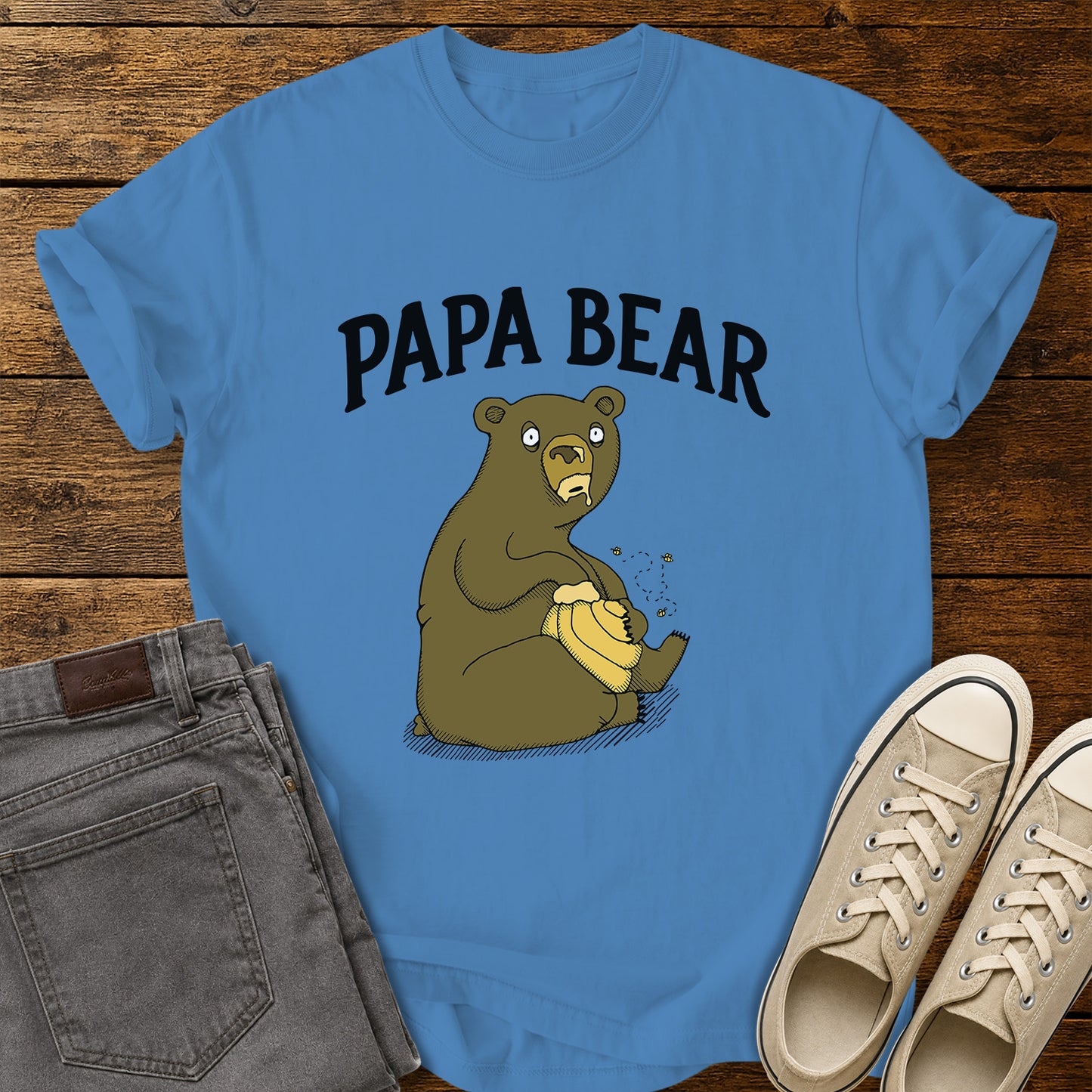 Royal blue flat-lay t-shirt mockup featuring a humorous cartoon bear sitting with a honey pot while bees buzz around it. Bold black lettering above the illustration reads, "PAPA BEAR." The vintage-inspired design is centered on the shirt, which is displayed on a rustic wood plank background alongside folded gray jeans and a pair of cream canvas sneakers.