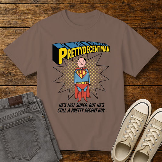 espresso brown t-shirt mockup featuring a comic-style “PRETTYDECENTMAN” superhero graphic with a hand-drawn character in a blue and red costume standing in front of a halftone starburst. Text below reads: “HE’S NOT SUPER, BUT HE’S STILL A PRETTY DECENT GUY.” Shirt is displayed on a rustic wood background with gray jeans and beige sneakers.