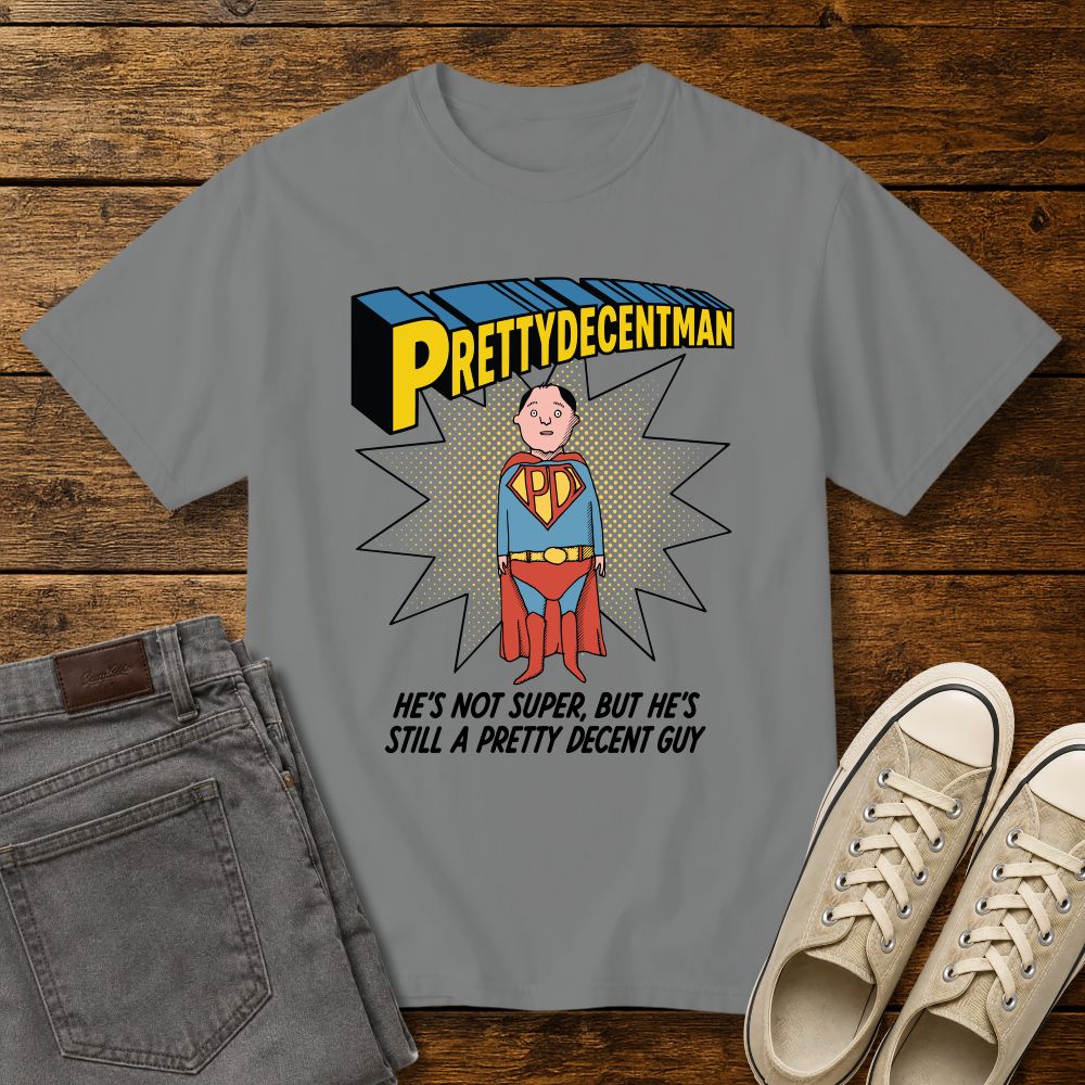grey t-shirt mockup featuring a comic-style “PRETTYDECENTMAN” superhero graphic with a hand-drawn character in a blue and red costume standing in front of a halftone starburst. Text below reads: “HE’S NOT SUPER, BUT HE’S STILL A PRETTY DECENT GUY.” Shirt is displayed on a rustic wood background with gray jeans and beige sneakers.