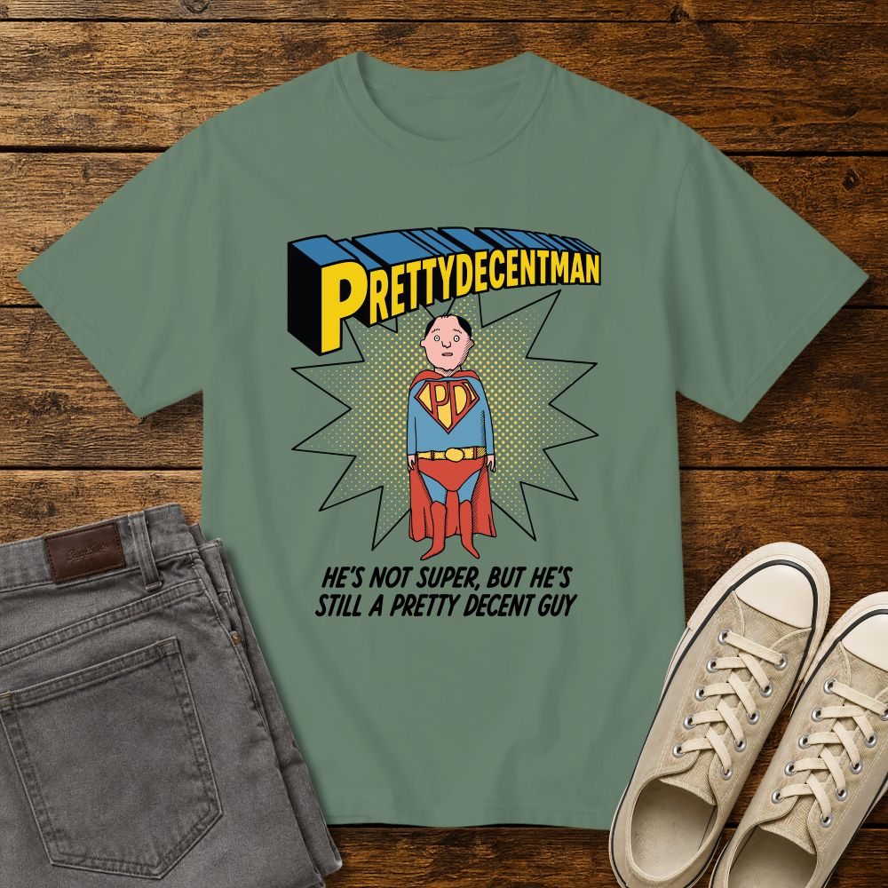 light green t-shirt mockup featuring a comic-style “PRETTYDECENTMAN” superhero graphic with a hand-drawn character in a blue and red costume standing in front of a halftone starburst. Text below reads: “HE’S NOT SUPER, BUT HE’S STILL A PRETTY DECENT GUY.” Shirt is displayed on a rustic wood background with gray jeans and beige sneakers.
