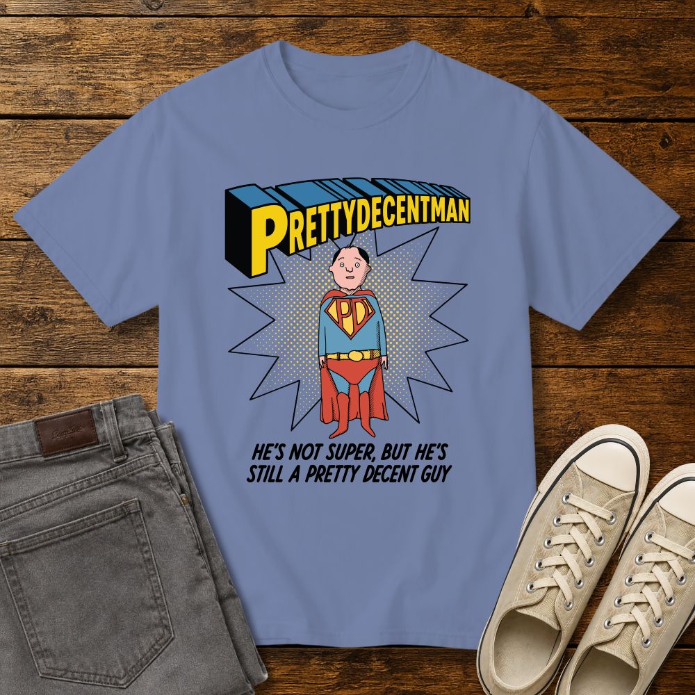washed denim color t-shirt mockup featuring a comic-style “PRETTYDECENTMAN” superhero graphic with a hand-drawn character in a blue and red costume standing in front of a halftone starburst. Text below reads: “HE’S NOT SUPER, BUT HE’S STILL A PRETTY DECENT GUY.” Shirt is displayed on a rustic wood background with gray jeans and beige sneakers.