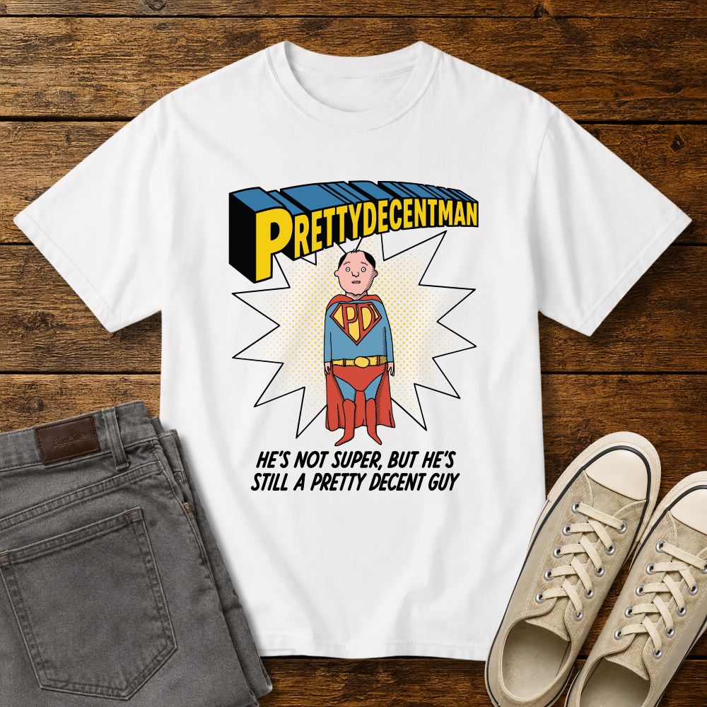 white t-shirt mockup featuring a comic-style “PRETTYDECENTMAN” superhero graphic with a hand-drawn character in a blue and red costume standing in front of a halftone starburst. Text below reads: “HE’S NOT SUPER, BUT HE’S STILL A PRETTY DECENT GUY.” Shirt is displayed on a rustic wood background with gray jeans and beige sneakers.