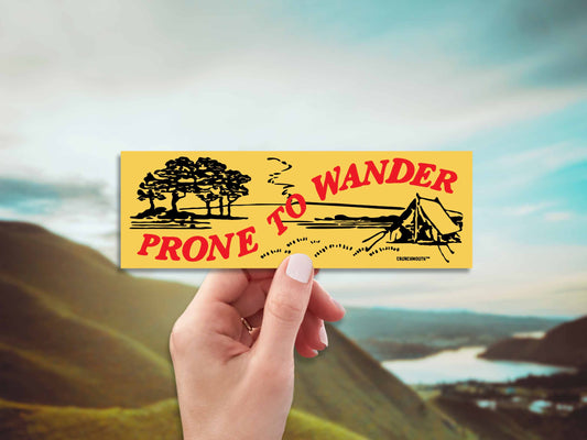 prone to wander camping bumper sticker, hand display, mountain skyline background